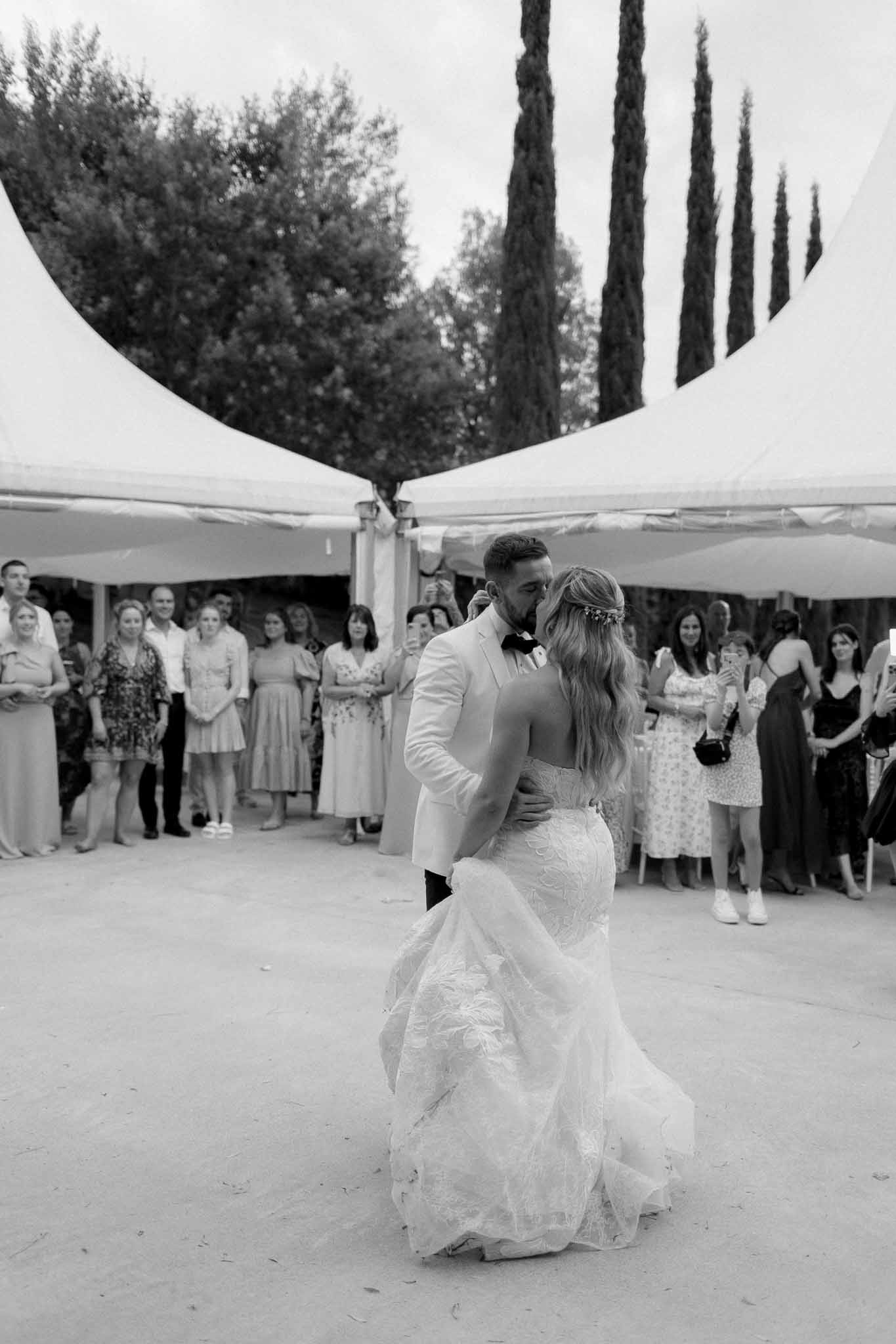 Black and white photo of couple kissing during first dance under white tent as guests watch at outdoor reception