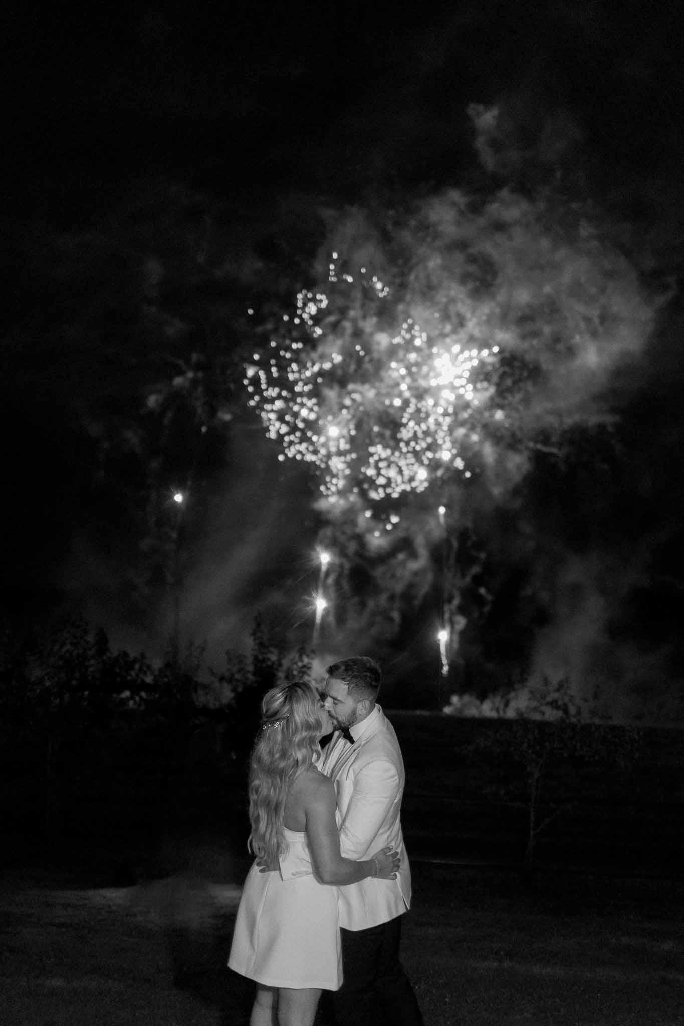 Bride and groom kissing outdoors at night with fireworks display behind them in black and white