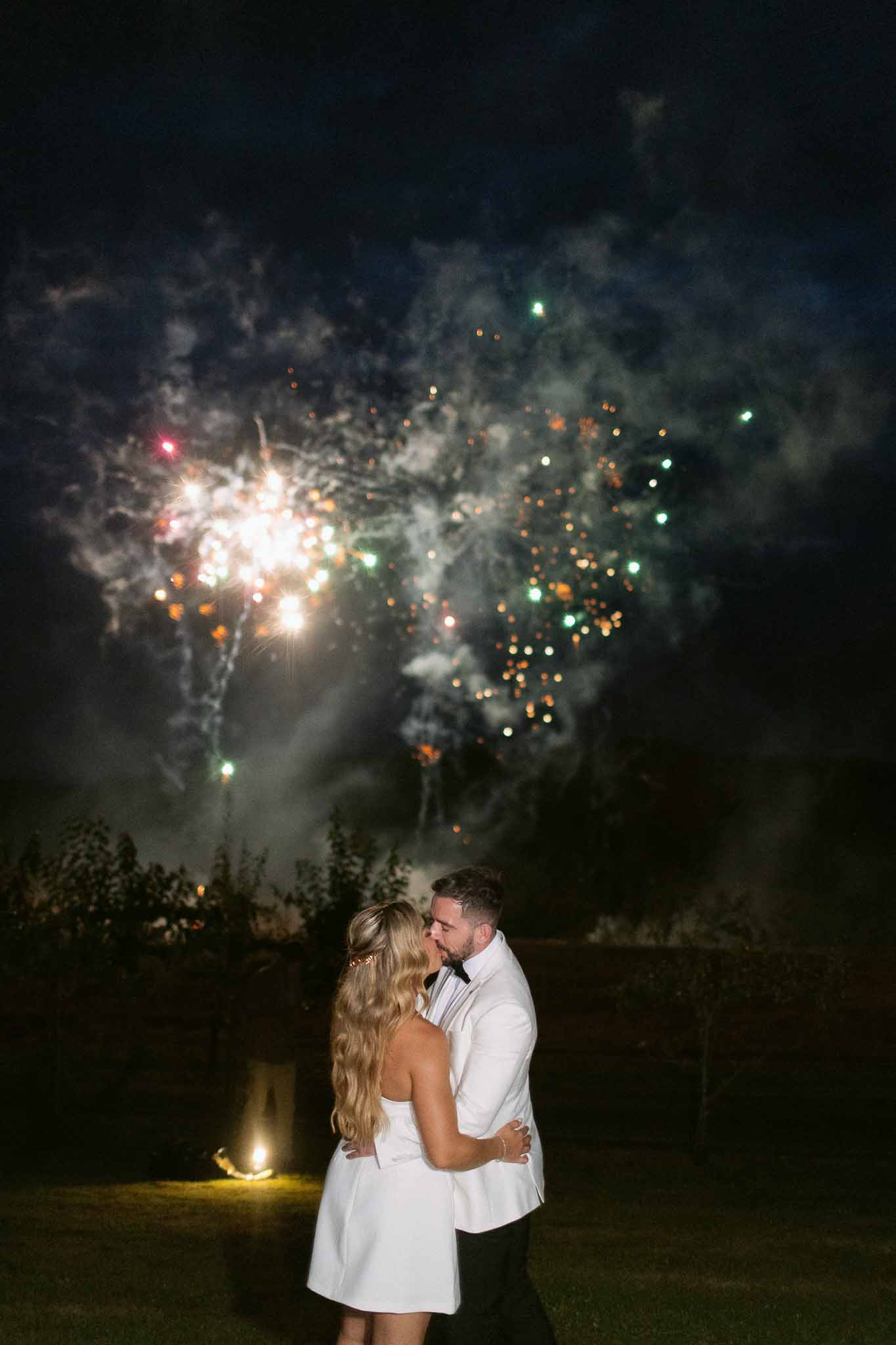 Bride in white mini dress and groom in white jacket kissing under multicolored fireworks display at night