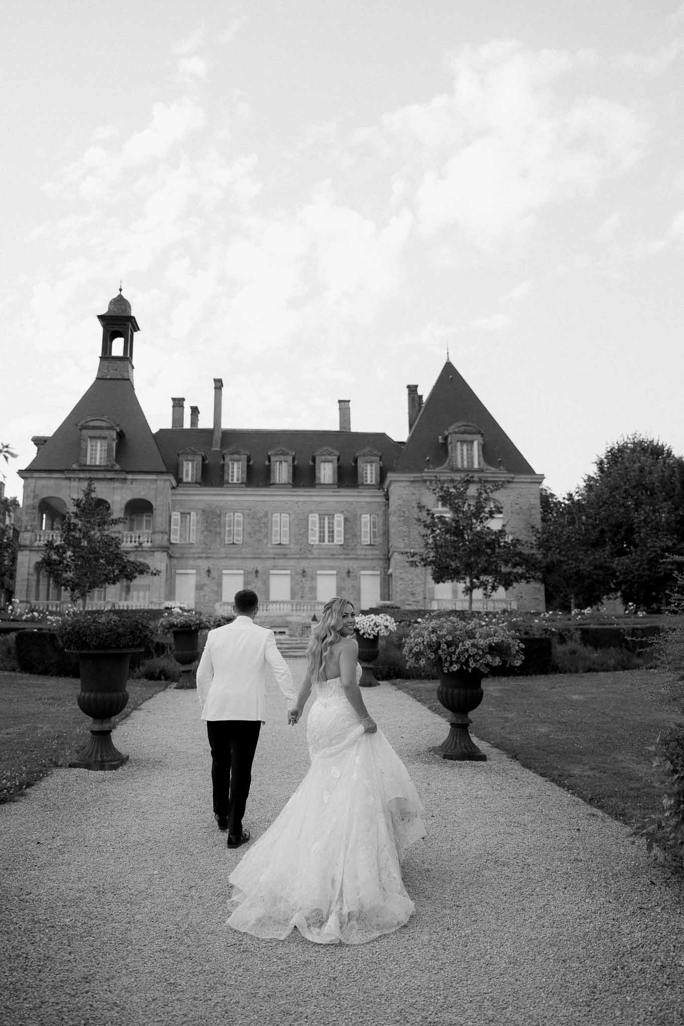 Couple walking toward grand château with manicured gardens and ornamental urns on wedding day