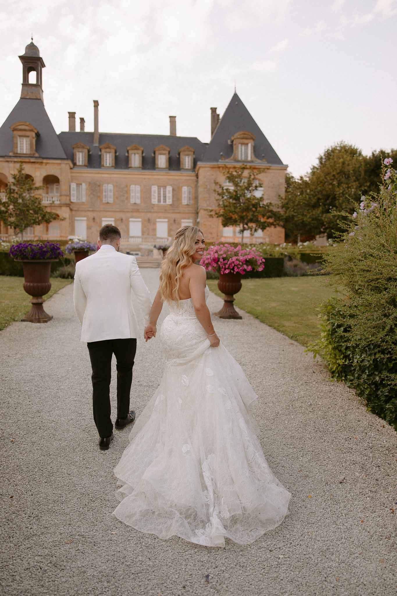 Bride and groom walking hand-in-hand toward grand brick château with formal gardens
