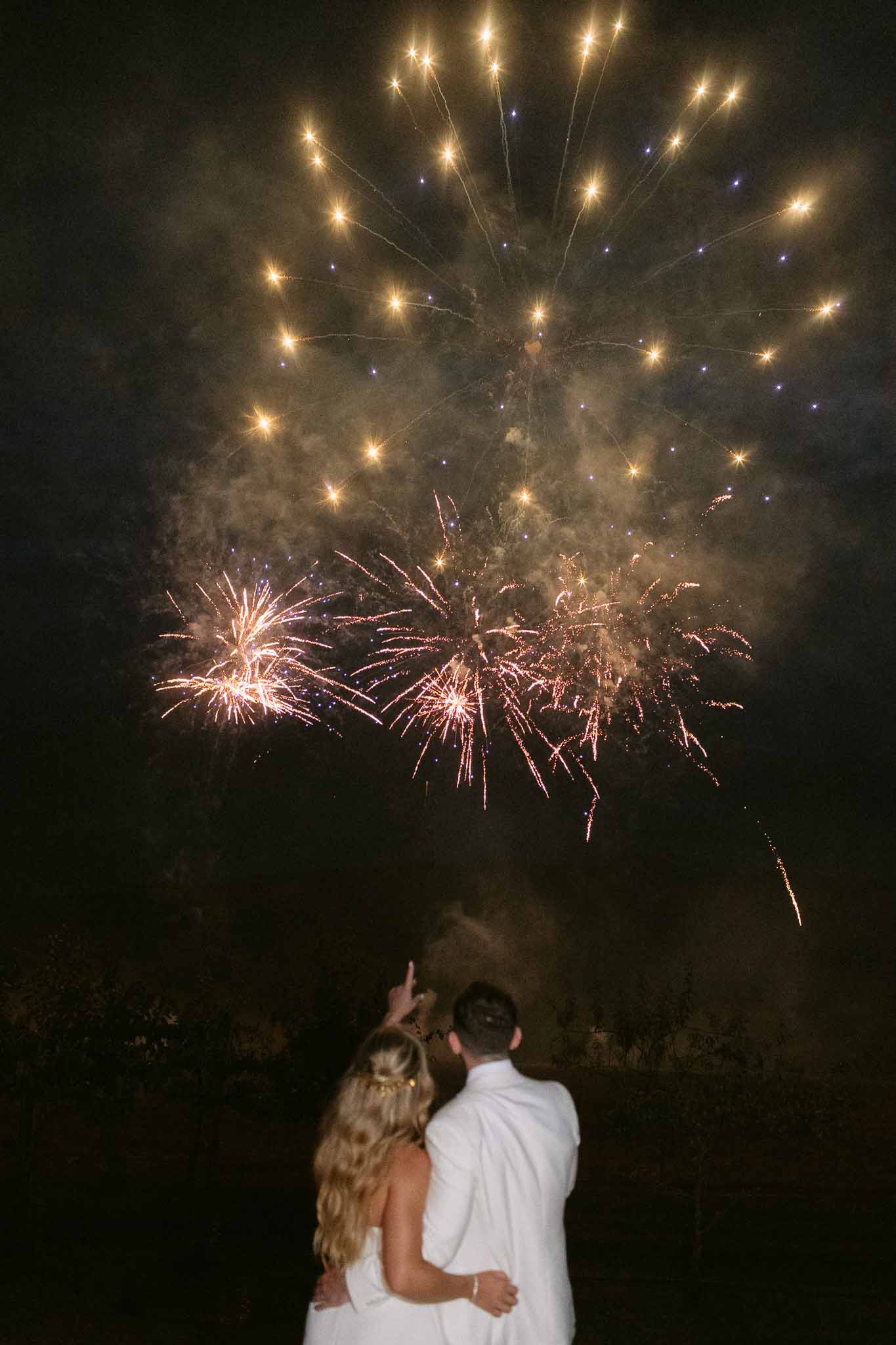 Bride and groom watching gold fireworks display from behind at night on venue grounds