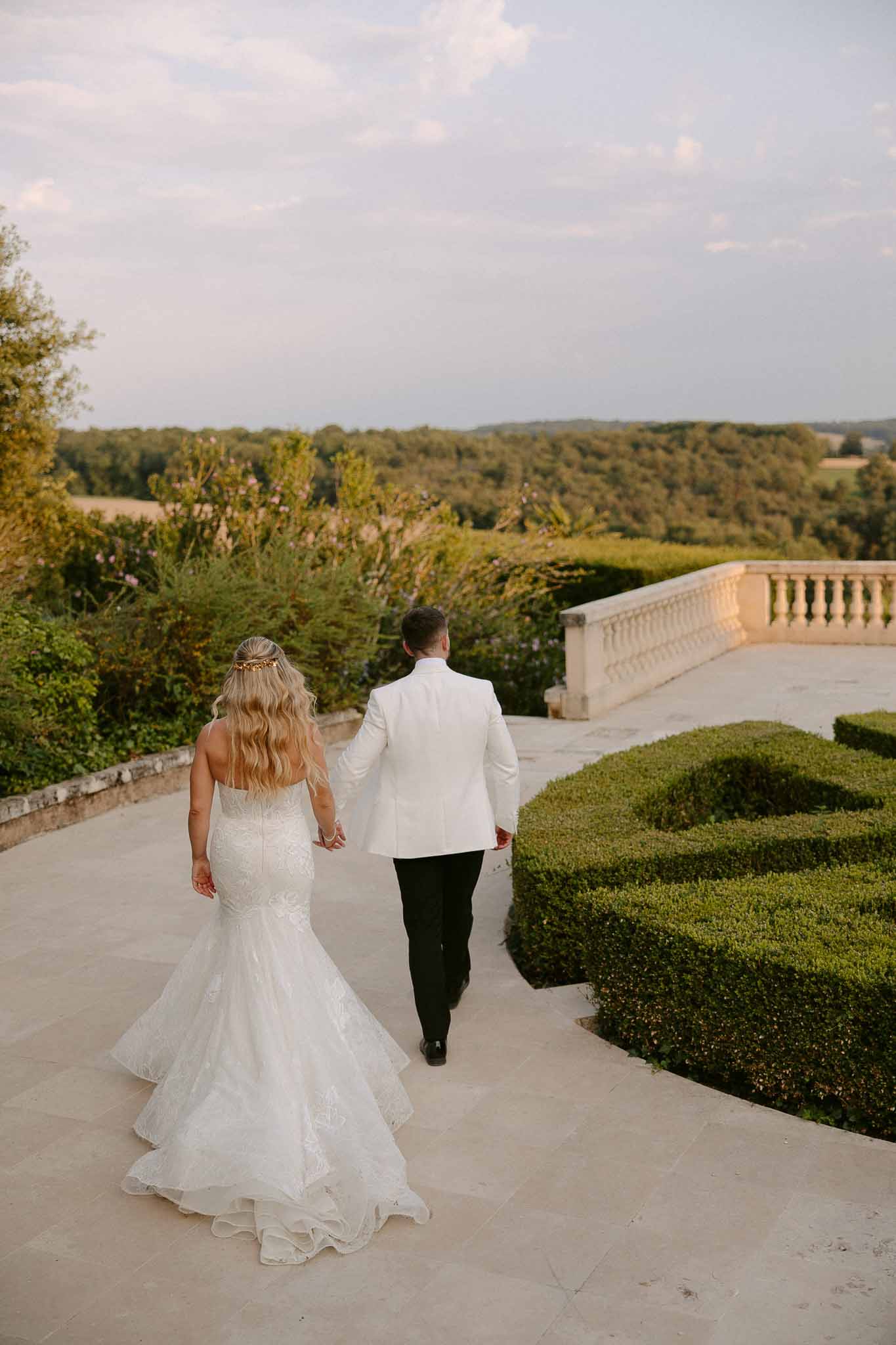 Bride and groom walking on stone terrace overlooking forested landscape at château estate