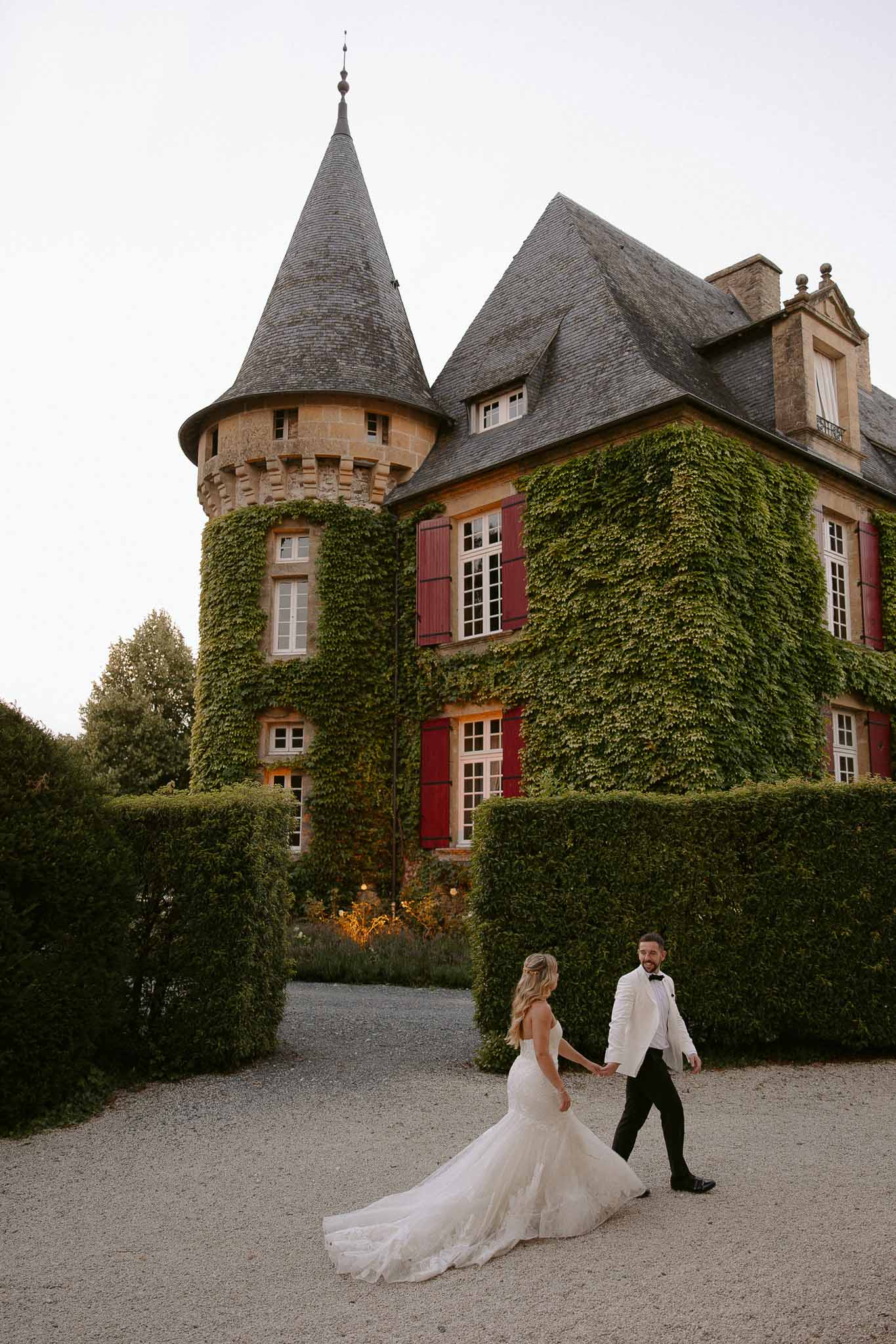 Bride and groom walking together on gravel courtyard path in front of château with conical tower