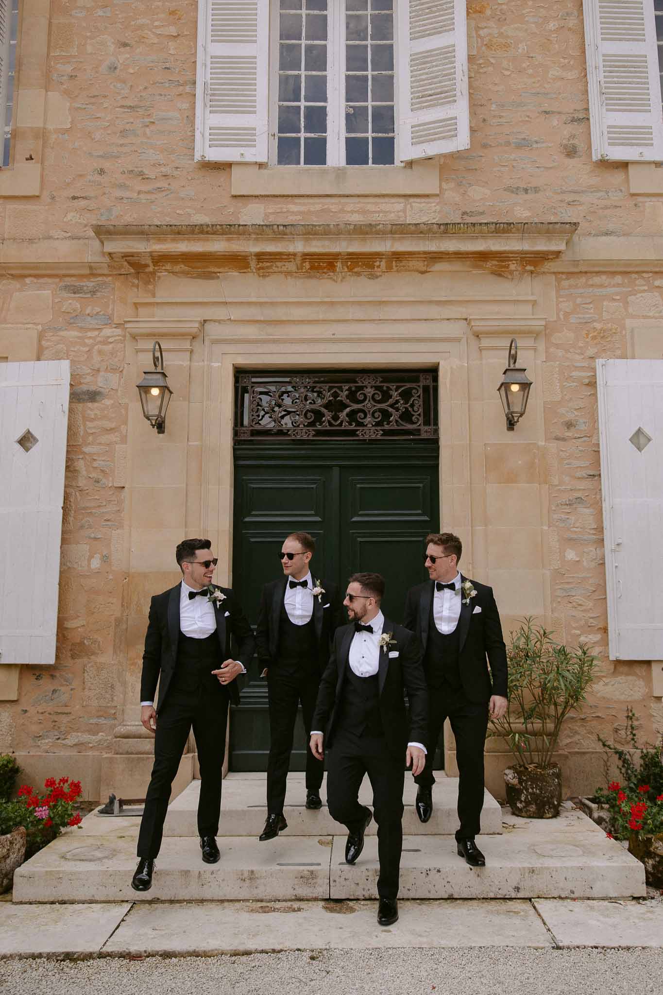 Four groomsmen in black tuxedos walking toward camera at stone courtyard entrance