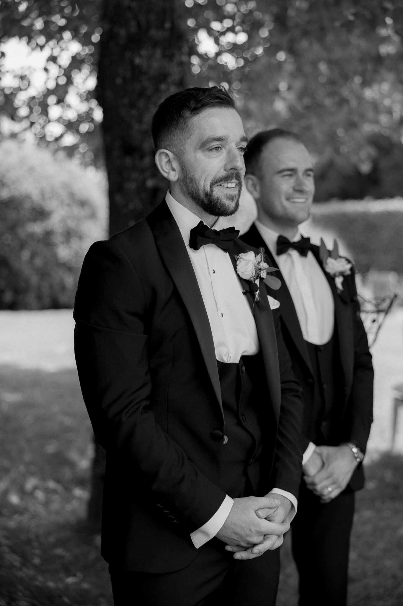 Groom and groomsman in black tuxedos during outdoor wedding ceremony with natural bokeh background