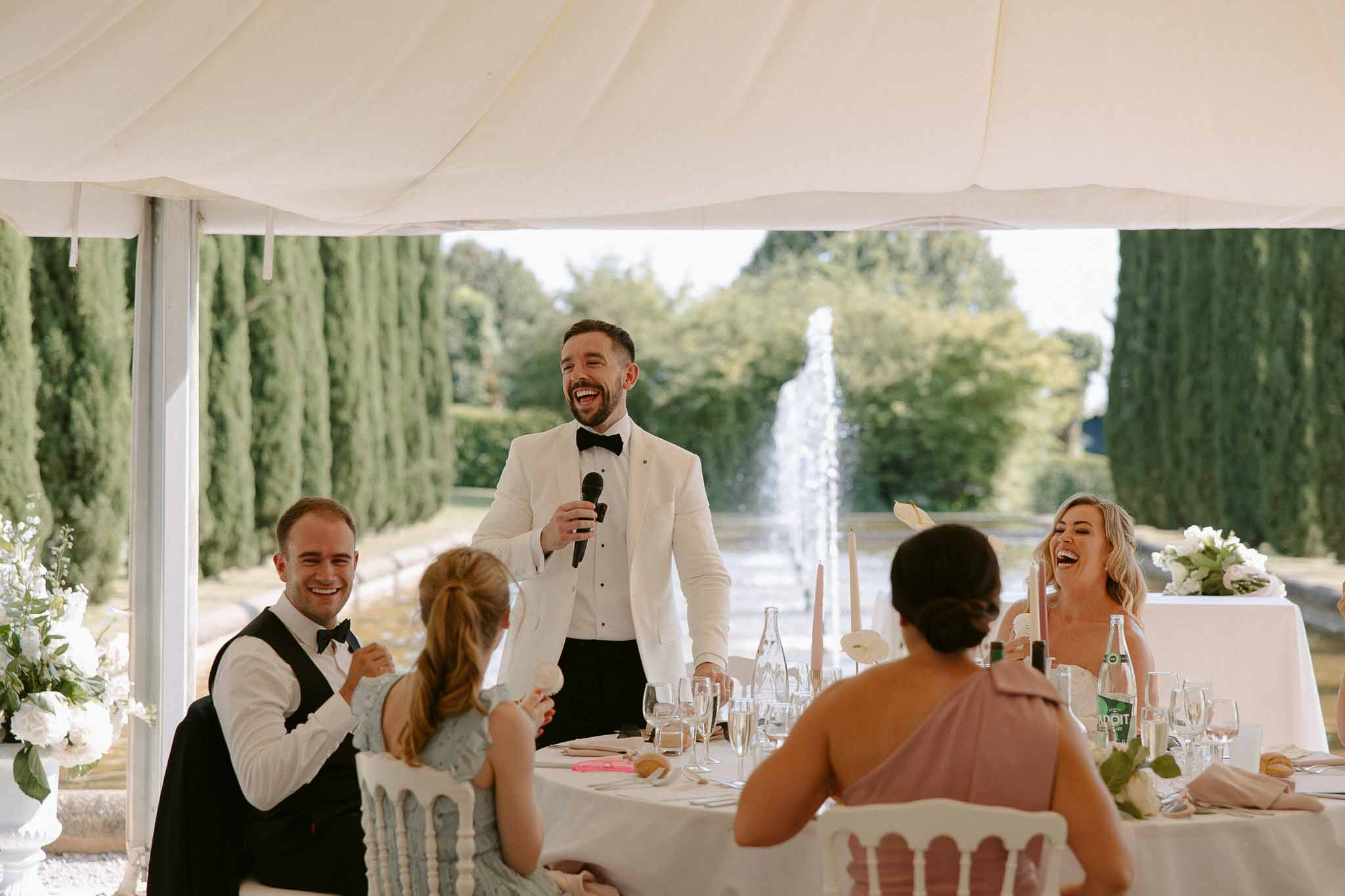 Groom delivering speech in white marquee as bride and guests laugh at table with white floral arrangements and candles