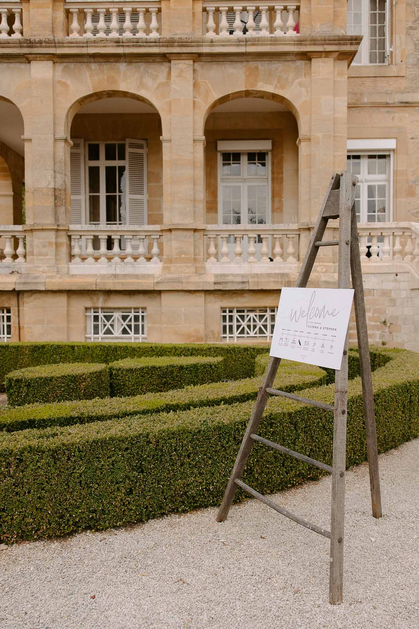 Minimalist welcome sign on rustic ladder easel before chateau facade with boxwood parterre garden