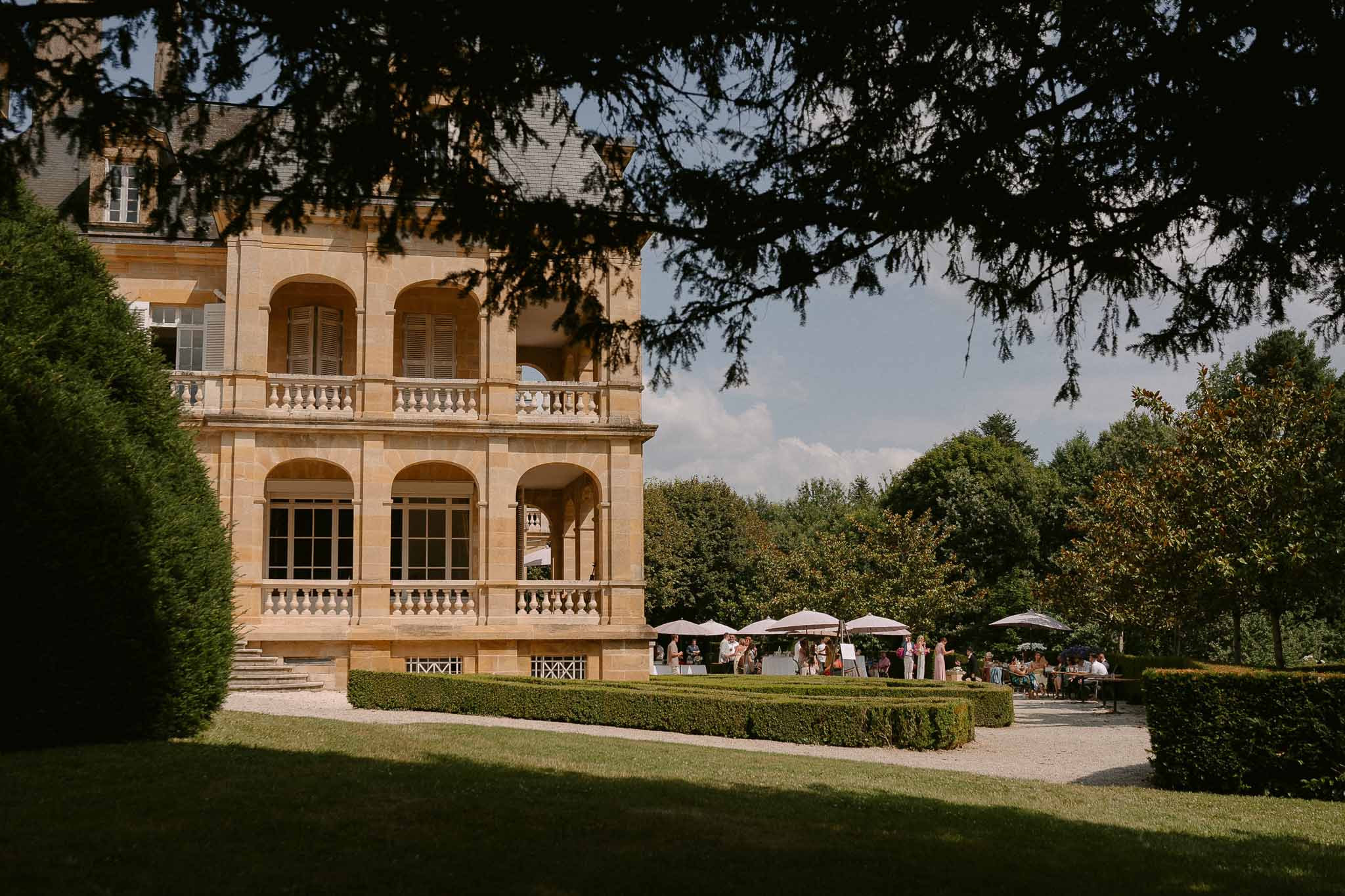 Guests under taupe umbrellas on gravel terrace beside golden limestone chateau with arched windows