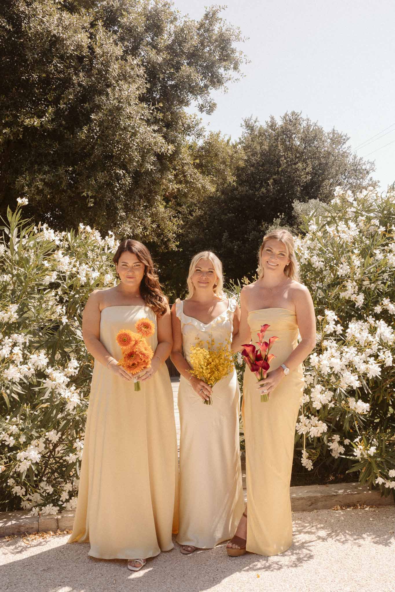 Bride and two bridesmaids holding colorful bouquets in Mediterranean garden with olive trees