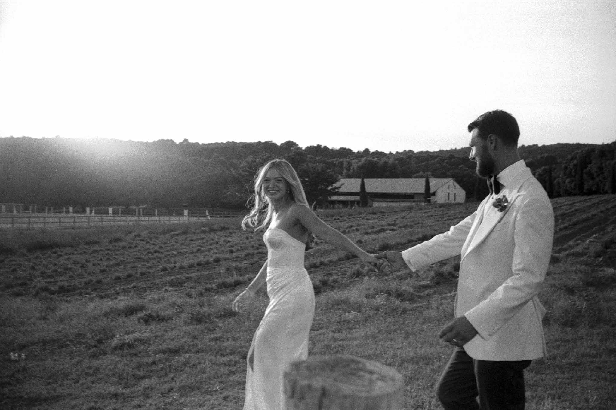 Bride and groom holding hands in countryside field with farm and hills in background