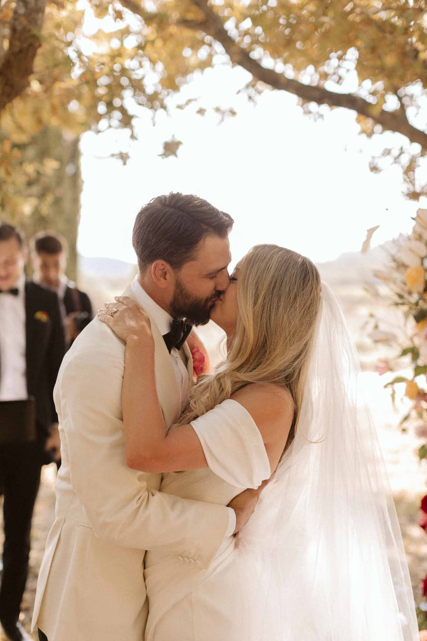 Couple's first kiss at outdoor ceremony under flowering arch at countryside wedding venue