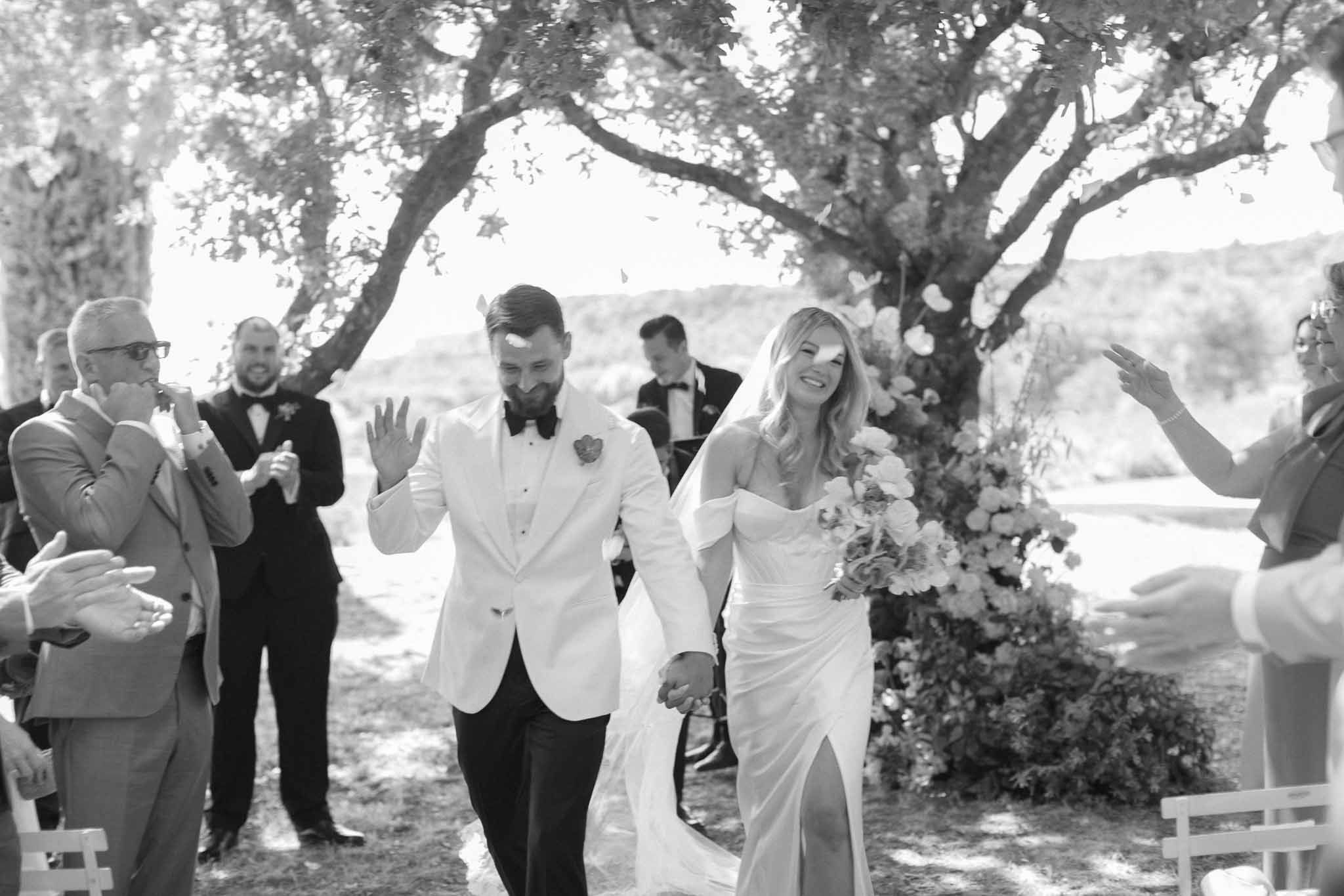 Newlywed couple walking down outdoor aisle after ceremony with guests applauding under large tree