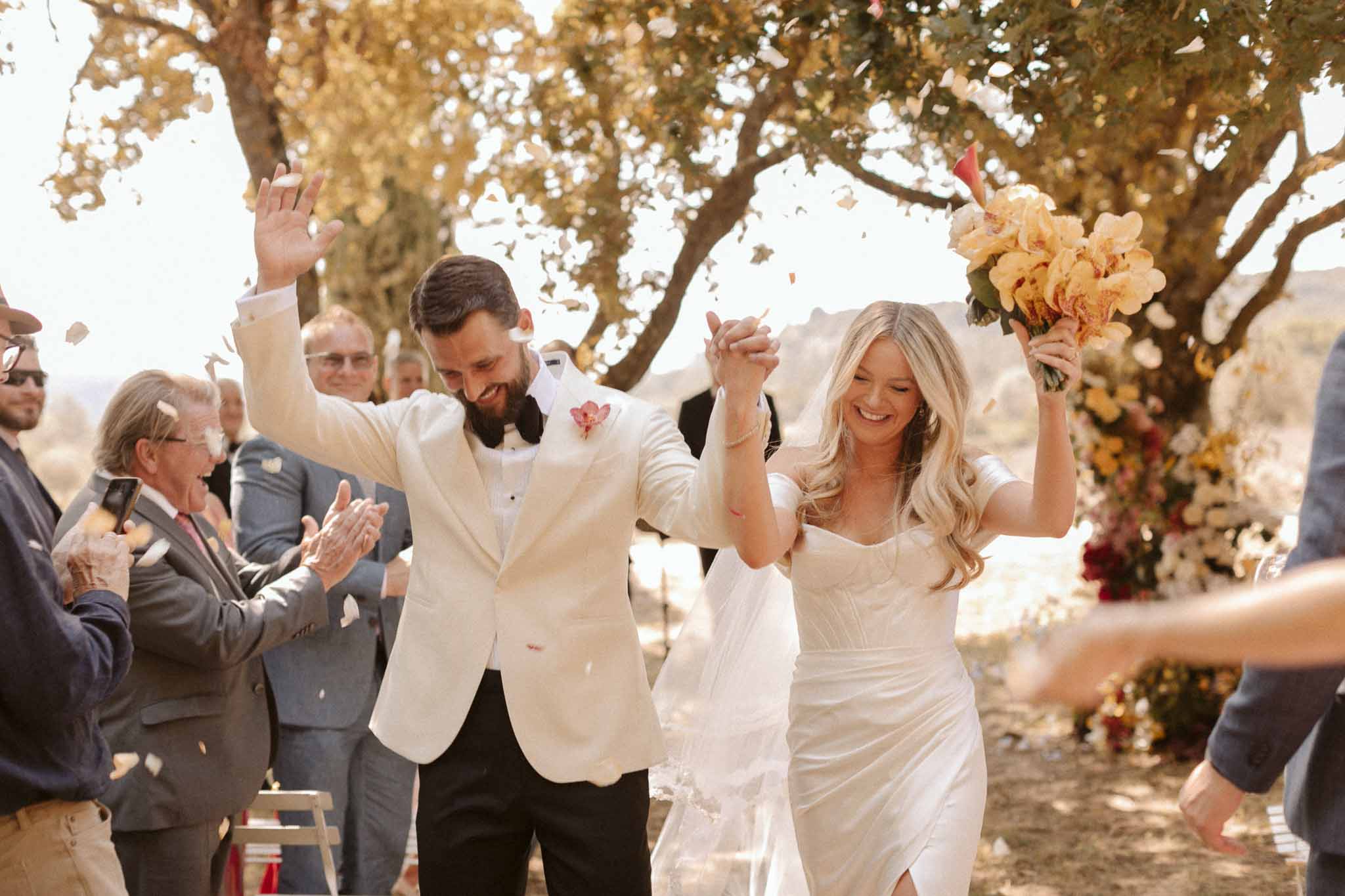 Bride and groom exit outdoor ceremony under tree as guests throw confetti at garden wedding venue