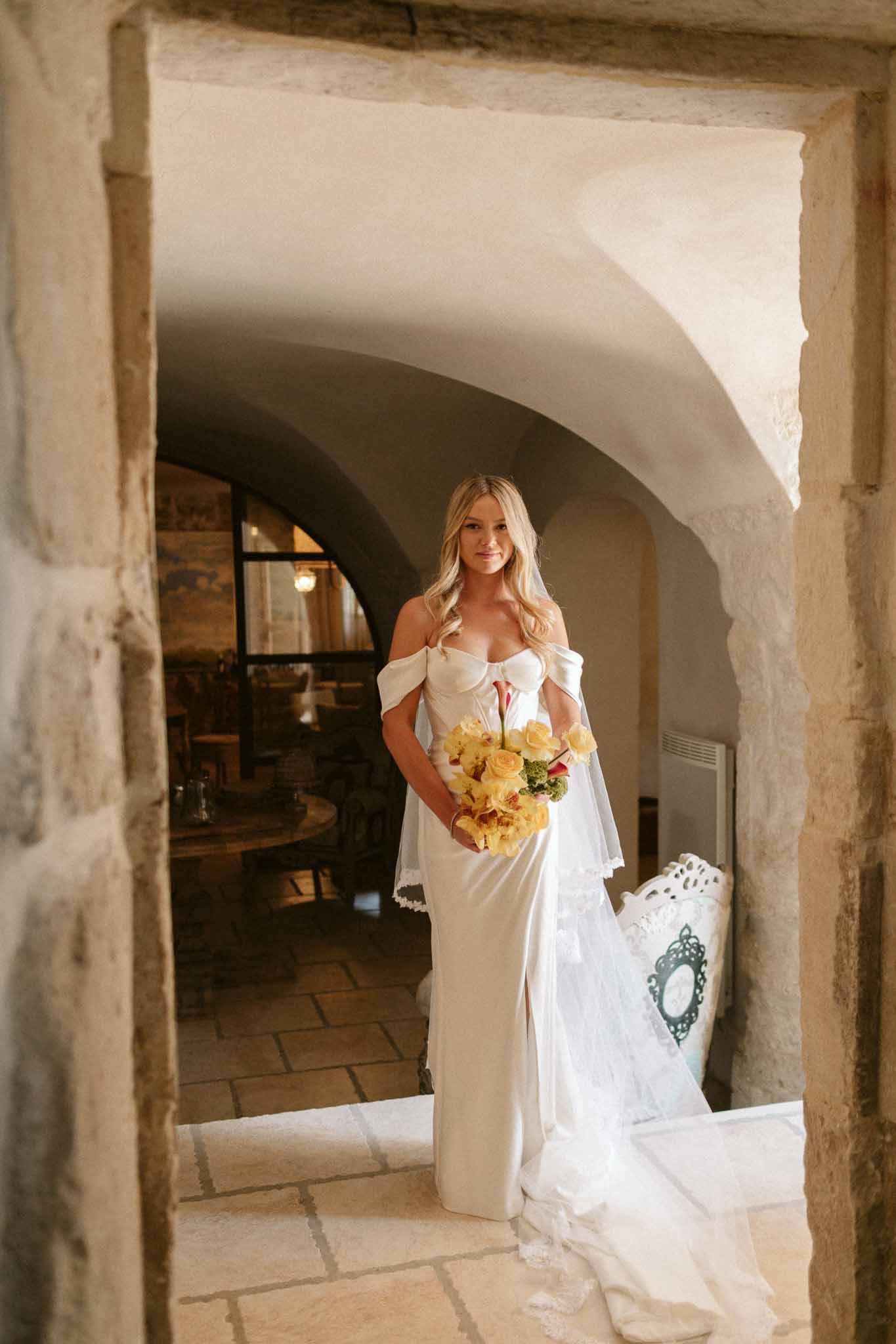 Bride in ivory dress with coral and yellow bouquet in stone archway at historic Mediterranean venue