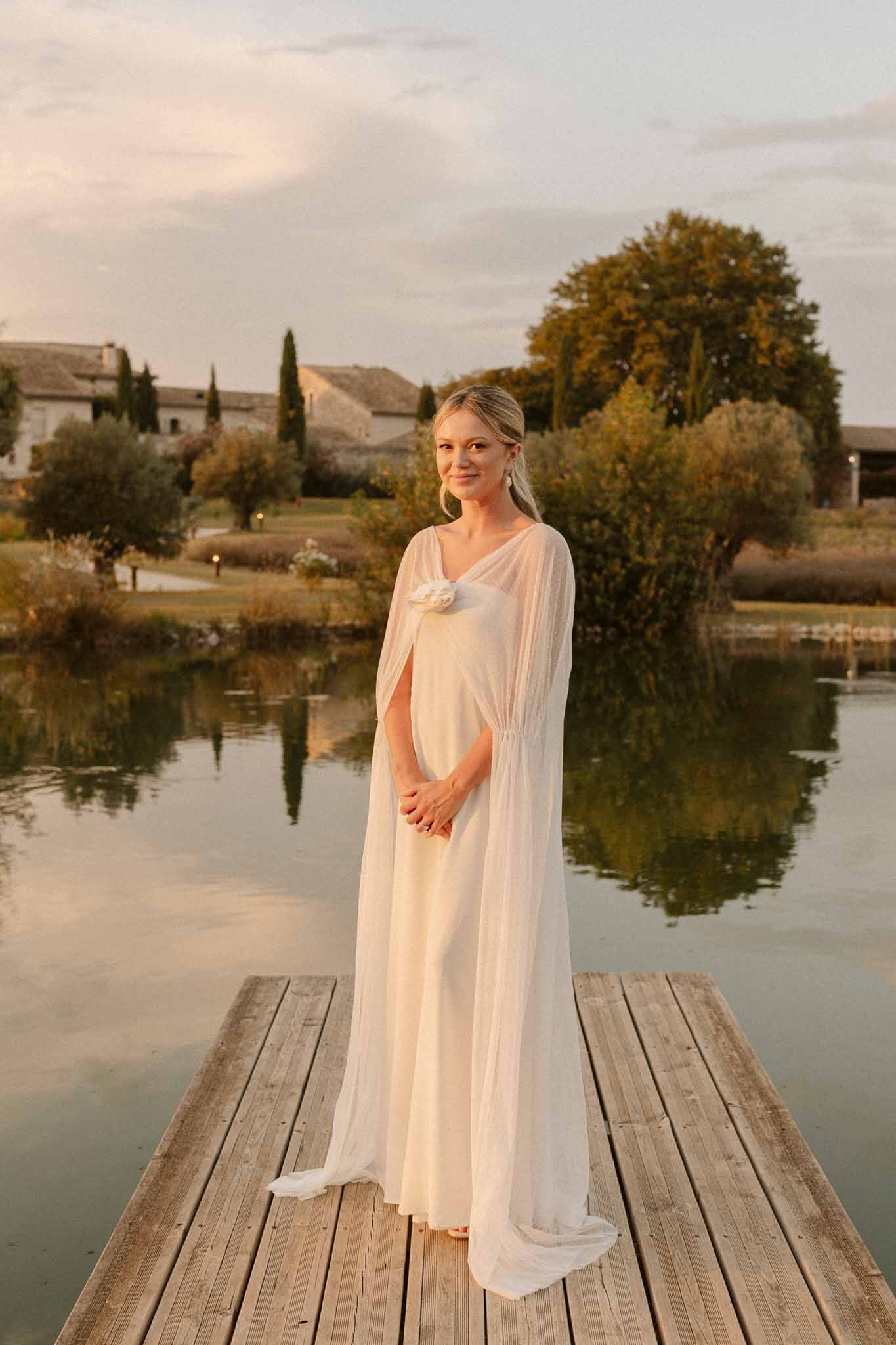 Bride in cream gown standing on wooden dock at Mediterranean estate with stone chapel in background