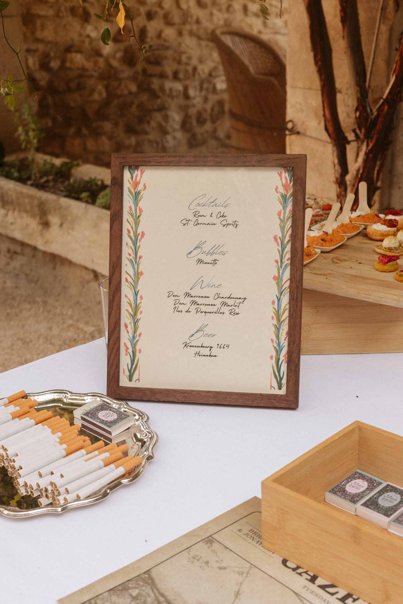 Hand-calligraphed cocktail menu board with floral details and wedding favors in rustic stone courtyard