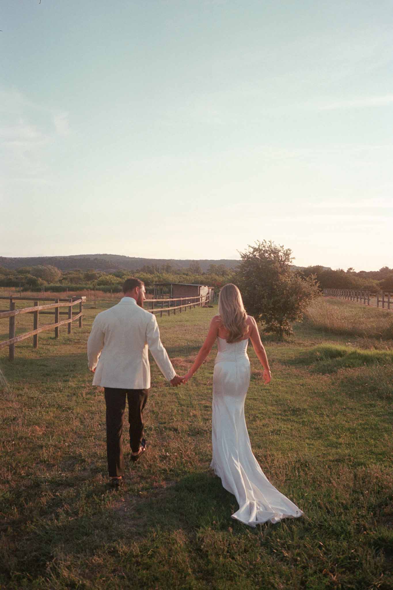 Bride and groom walking hand-in-hand across rural field at ranch wedding venue during golden hour