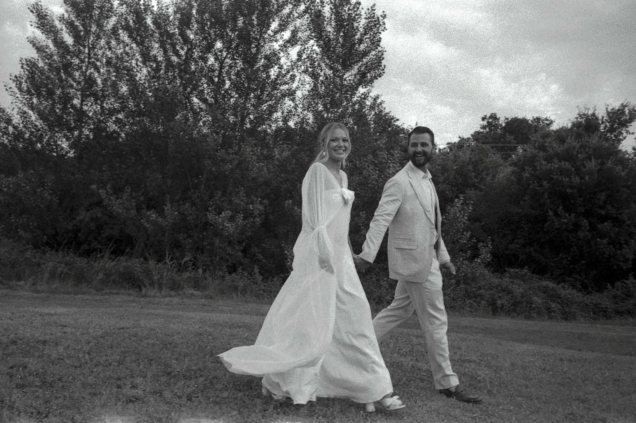 Bride and groom walking hand in hand through field with poplar trees in black and white portrait