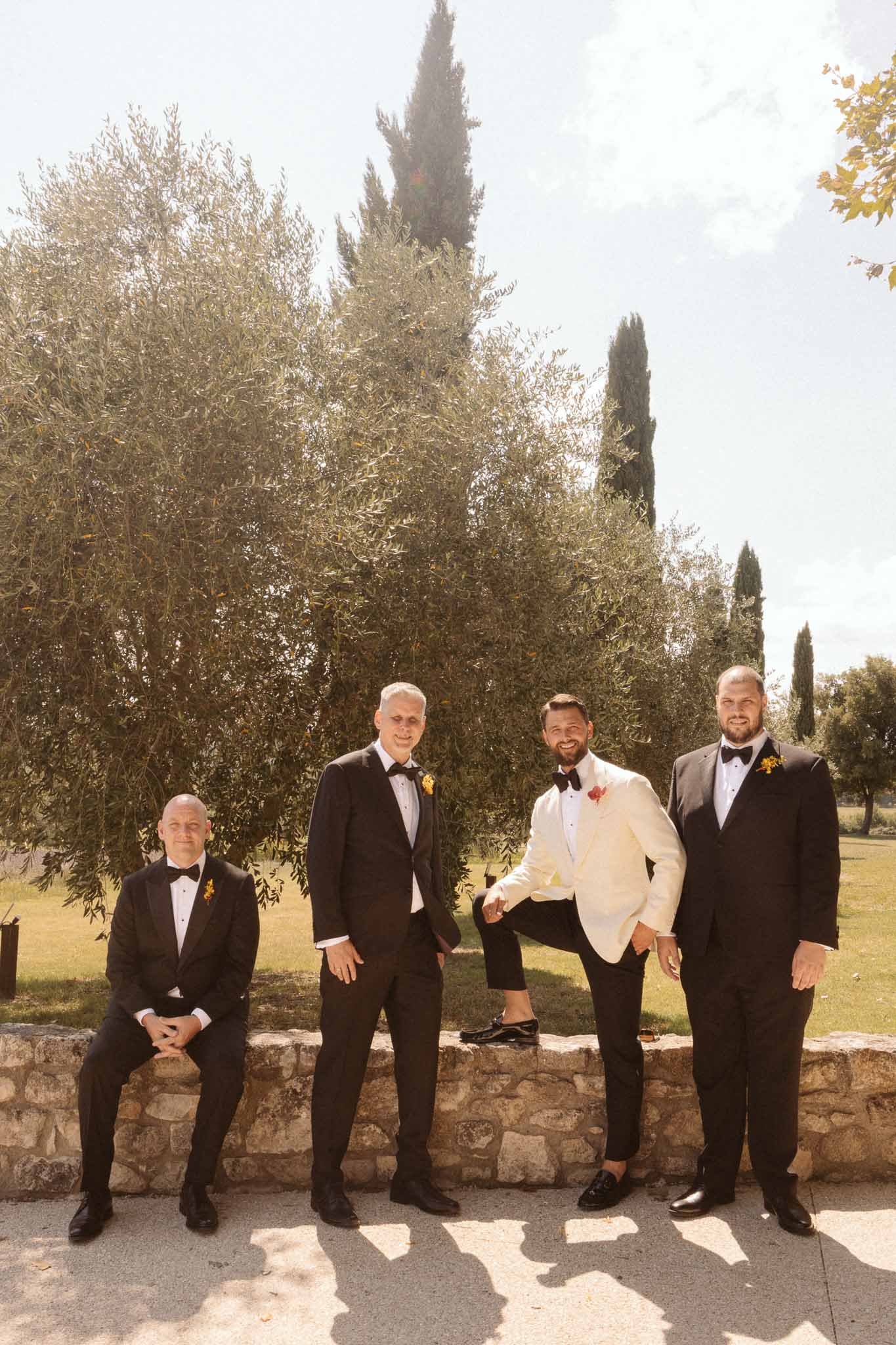 Groom and three groomsmen in formal attire posing outdoors among cypress and olive trees