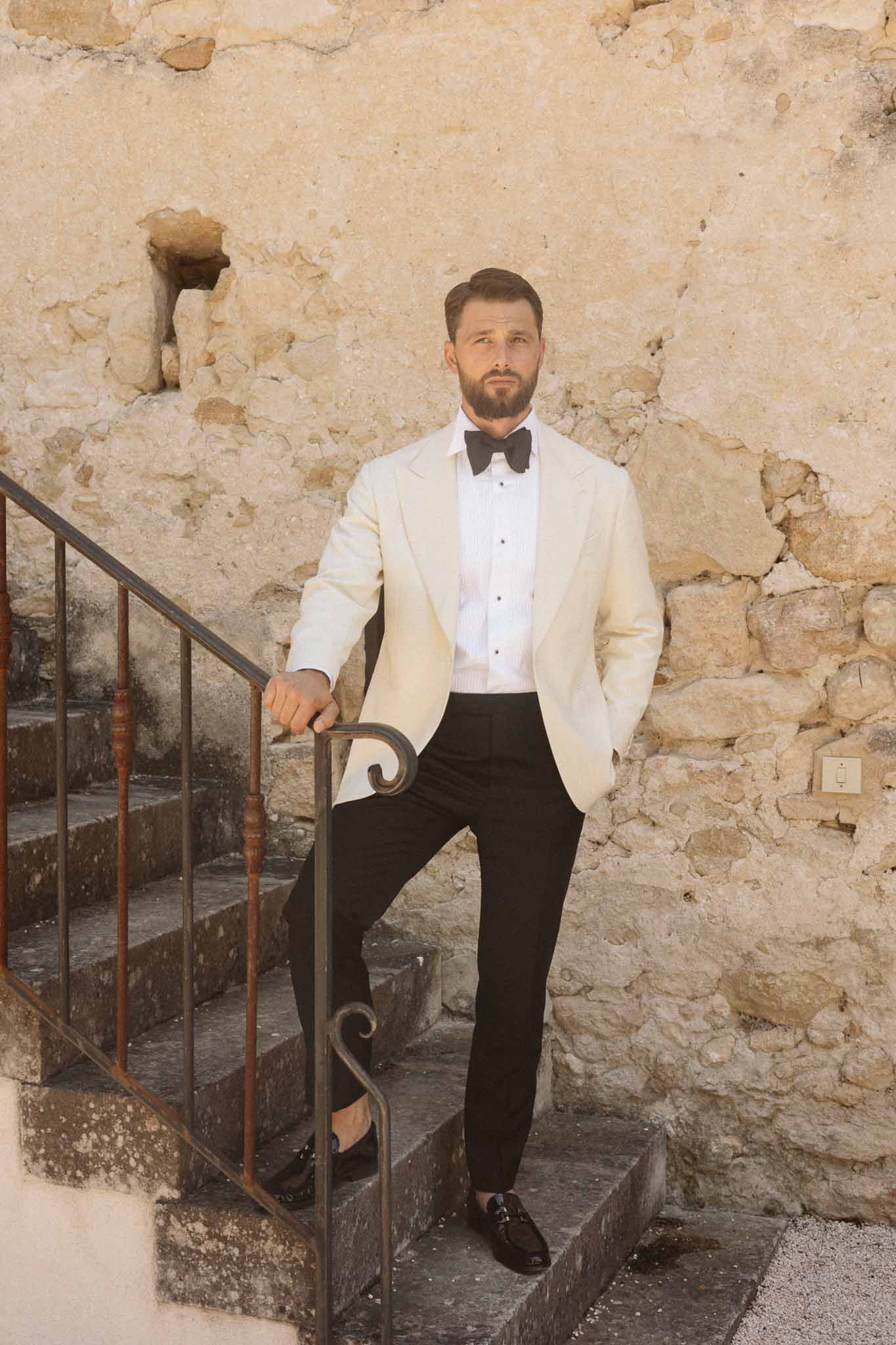 Groom in cream blazer and black bow tie posing on stone staircase at historic venue