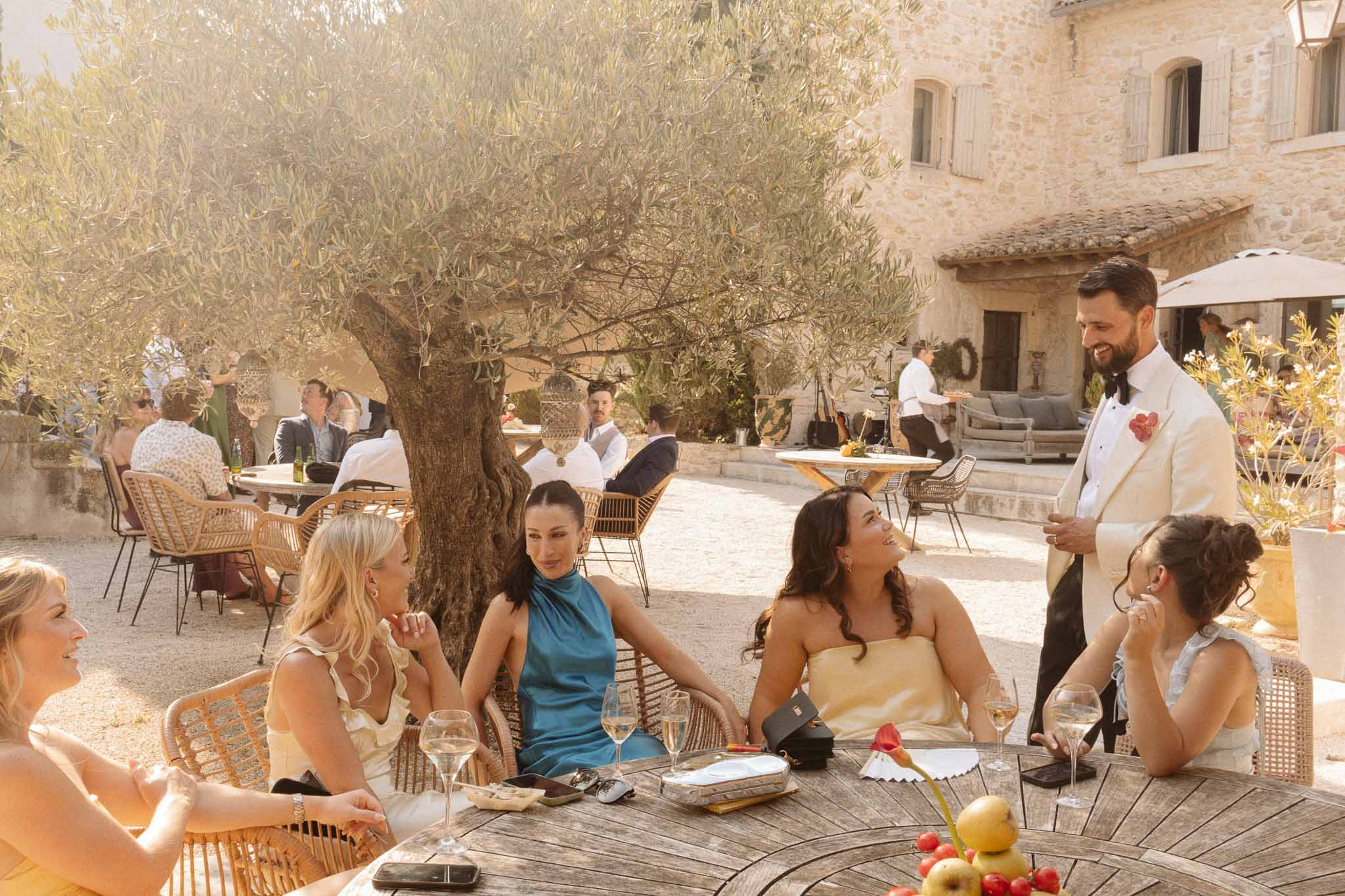 Reception gathering with guests mingling around olive tree in Tuscan stone courtyard with dining tables
