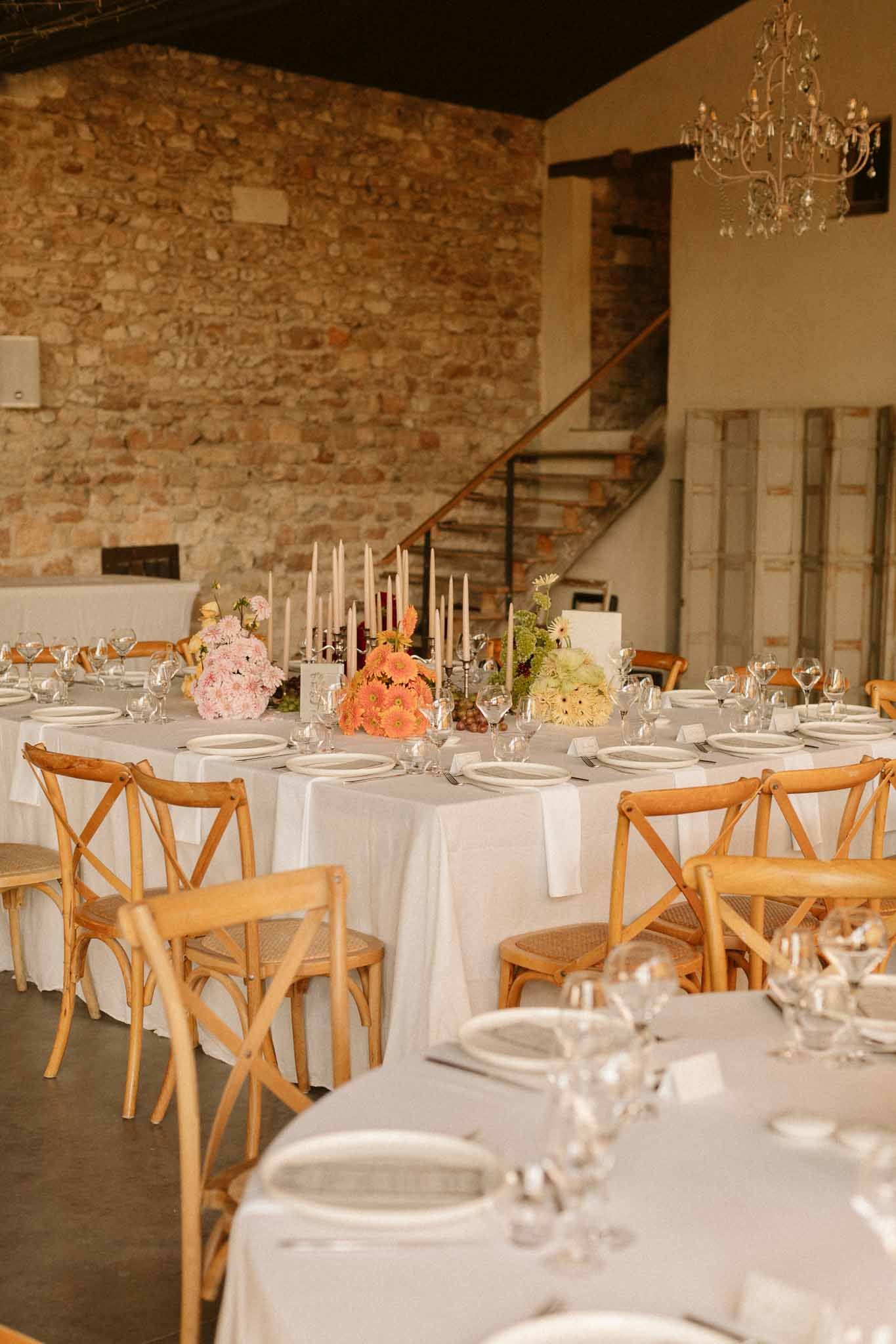 Indoor reception table setting with ivory linens and blush florals in stone-walled venue with chandelier
