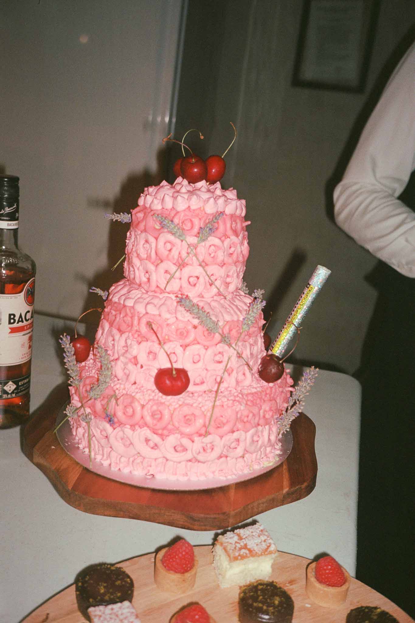 Three-tiered wedding cake with pink buttercream frosting and cherry decorations with petit four samples