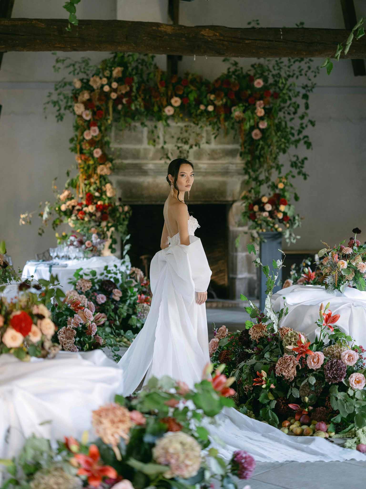 Bride in ivory gown poses beside elaborate floral installation in indoor reception space with exposed beams