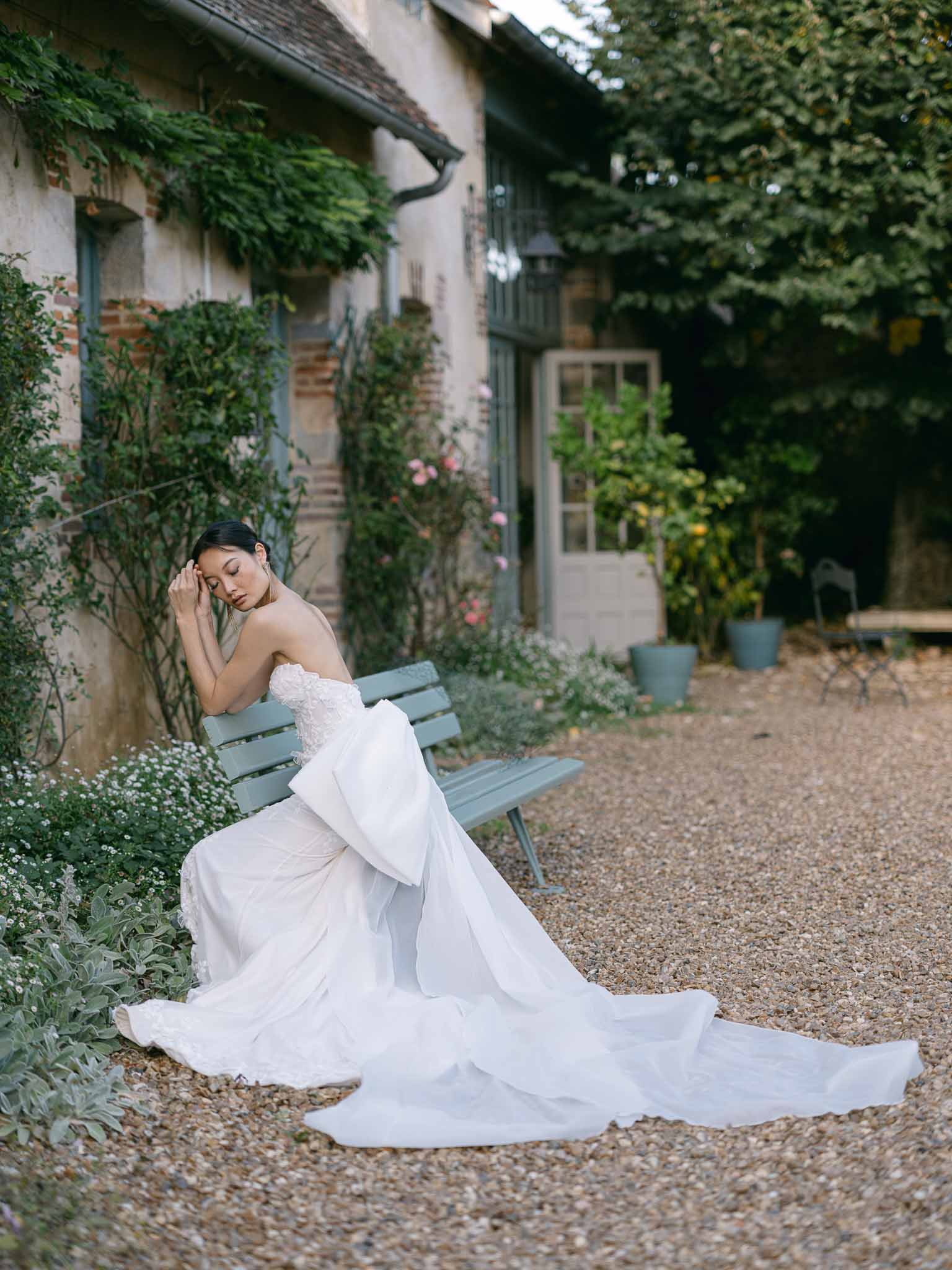 Bride in white gown sitting contemplatively on bench in ivy-covered garden courtyard
