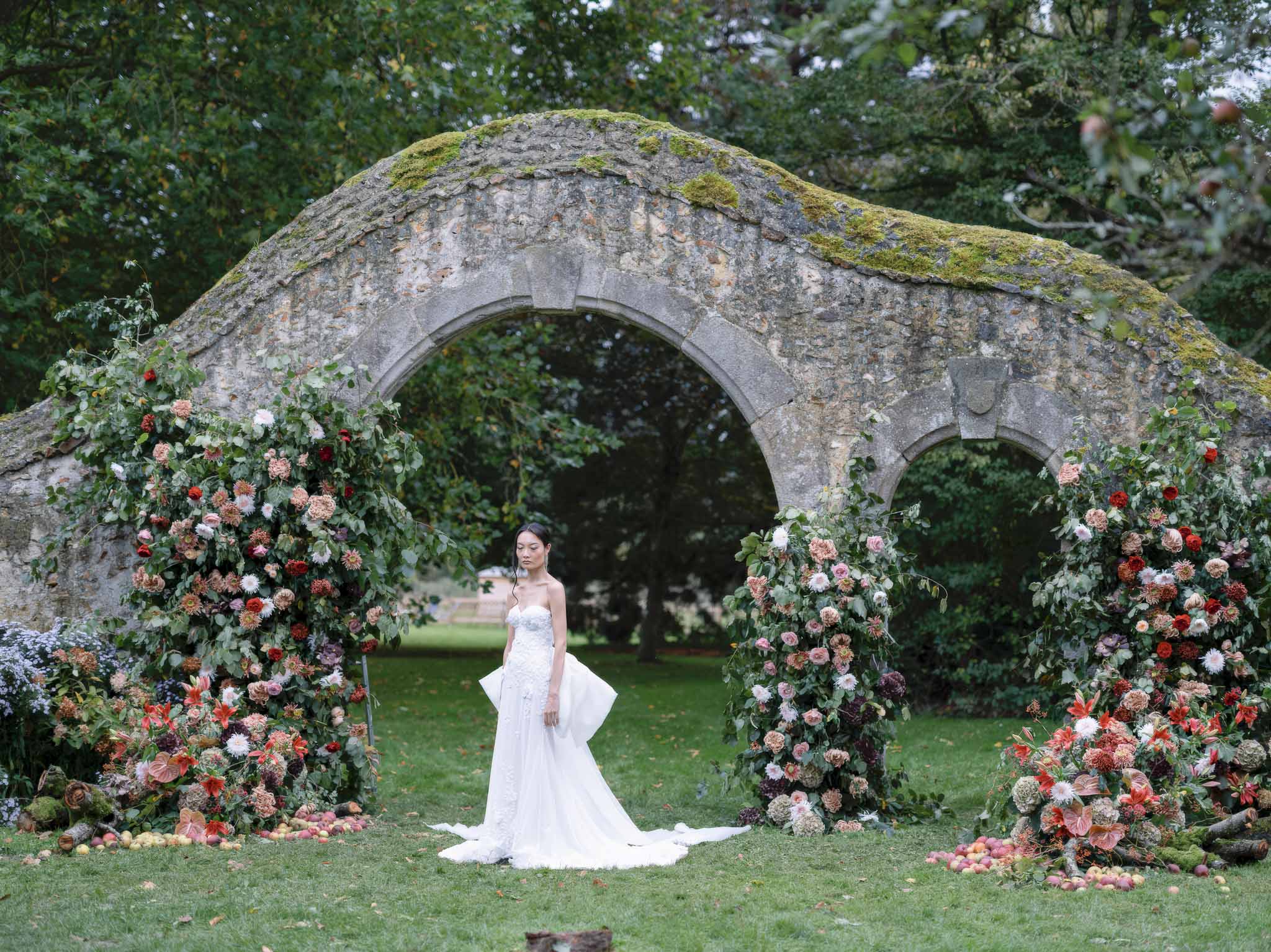 Bride in white wedding dress standing under stone archway with floral arrangements in garden setting
