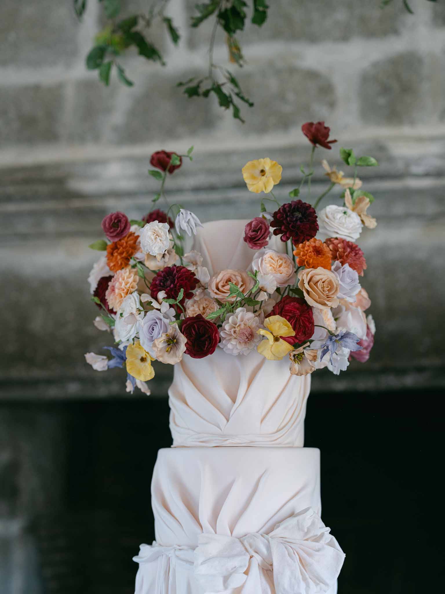 Two-tier wedding cake with mixed floral arrangement against stone wall backdrop