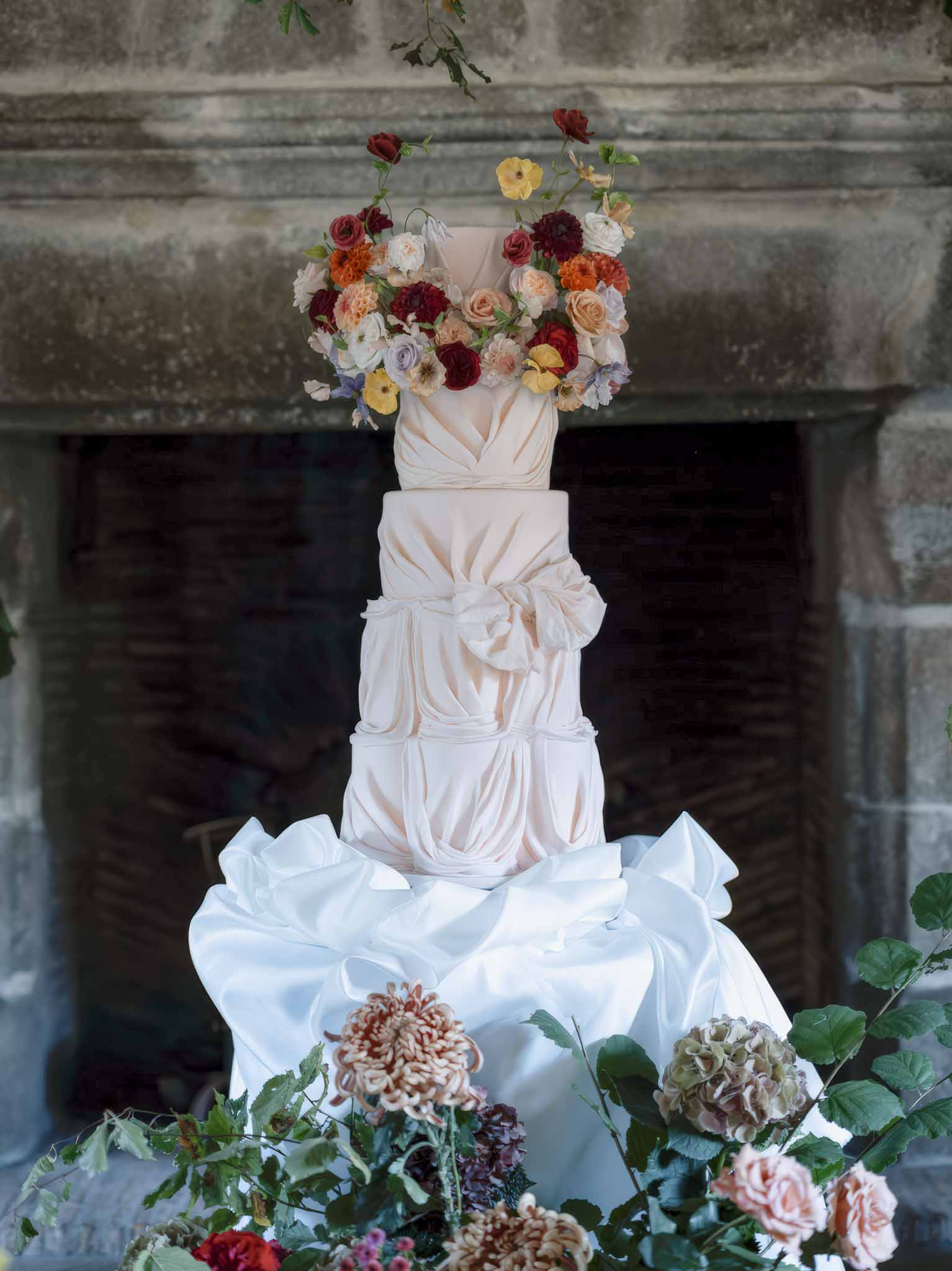 Four-tiered ivory wedding cake with burgundy and peach florals against stone archway