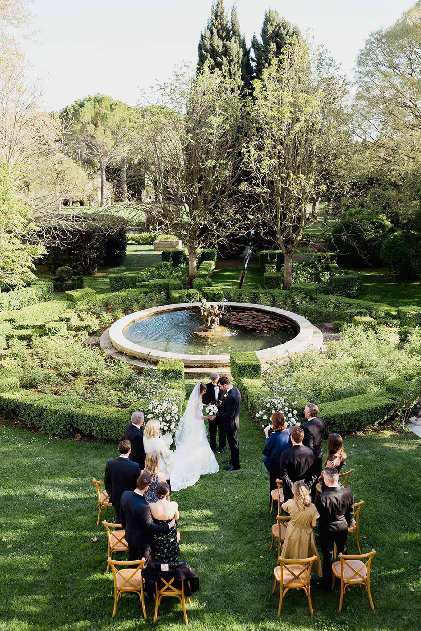 Outdoor wedding ceremony with guests in formal garden setting with fountain and cypress trees