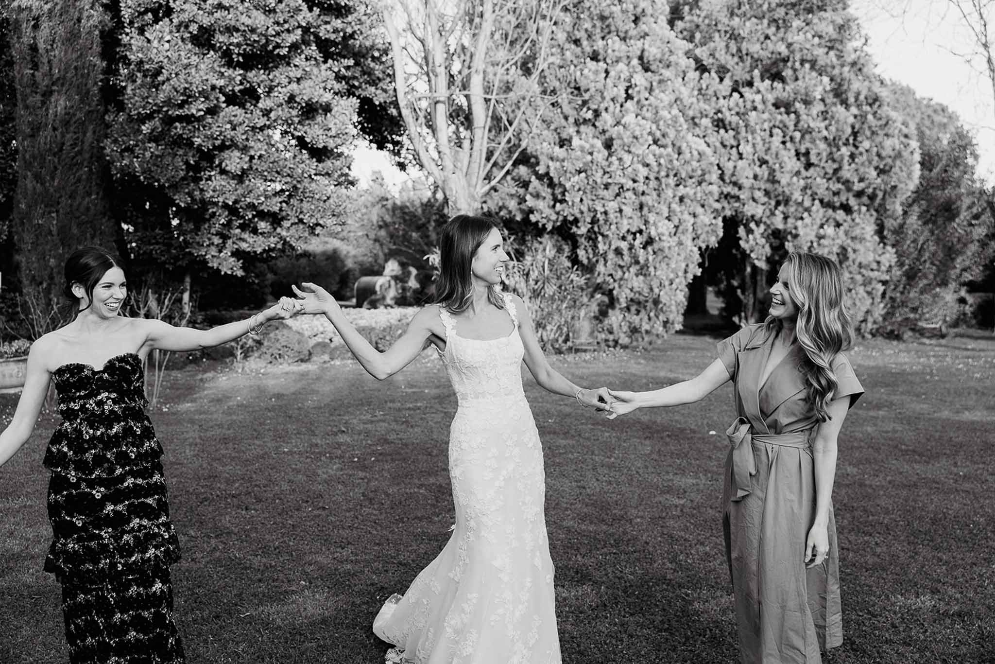 Bride with two attendants in formal dresses standing in manicured garden with hedges and trees