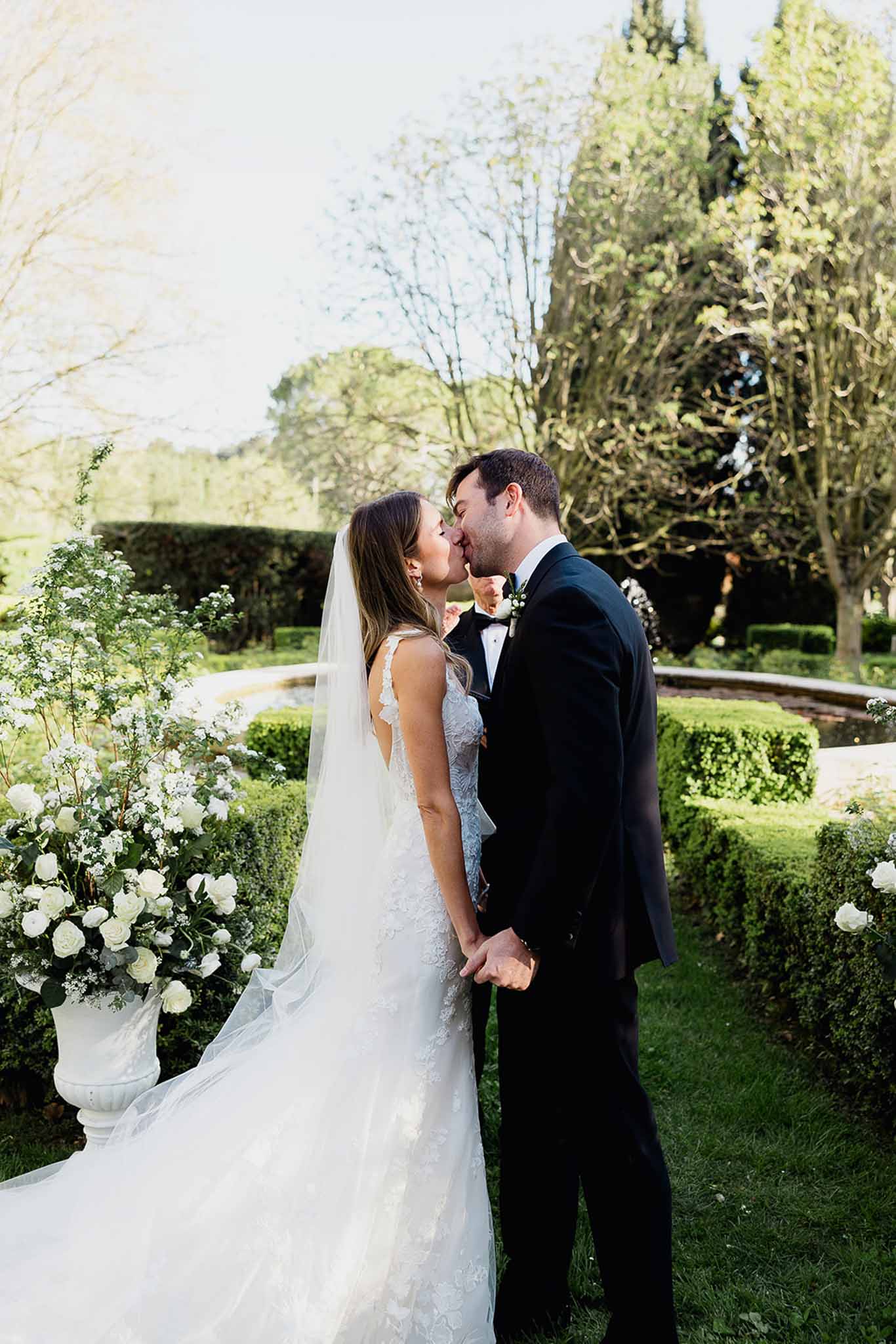 Bride and groom kissing in formal garden with white roses and classical urn