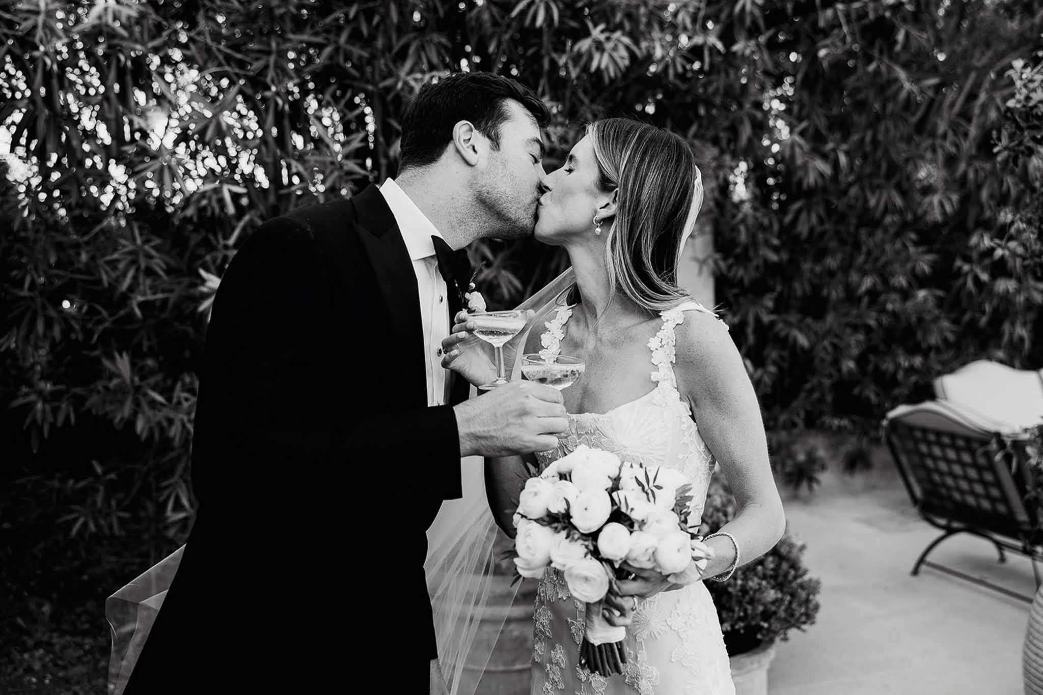 Bride and groom kissing with champagne flutes in garden setting with string lights