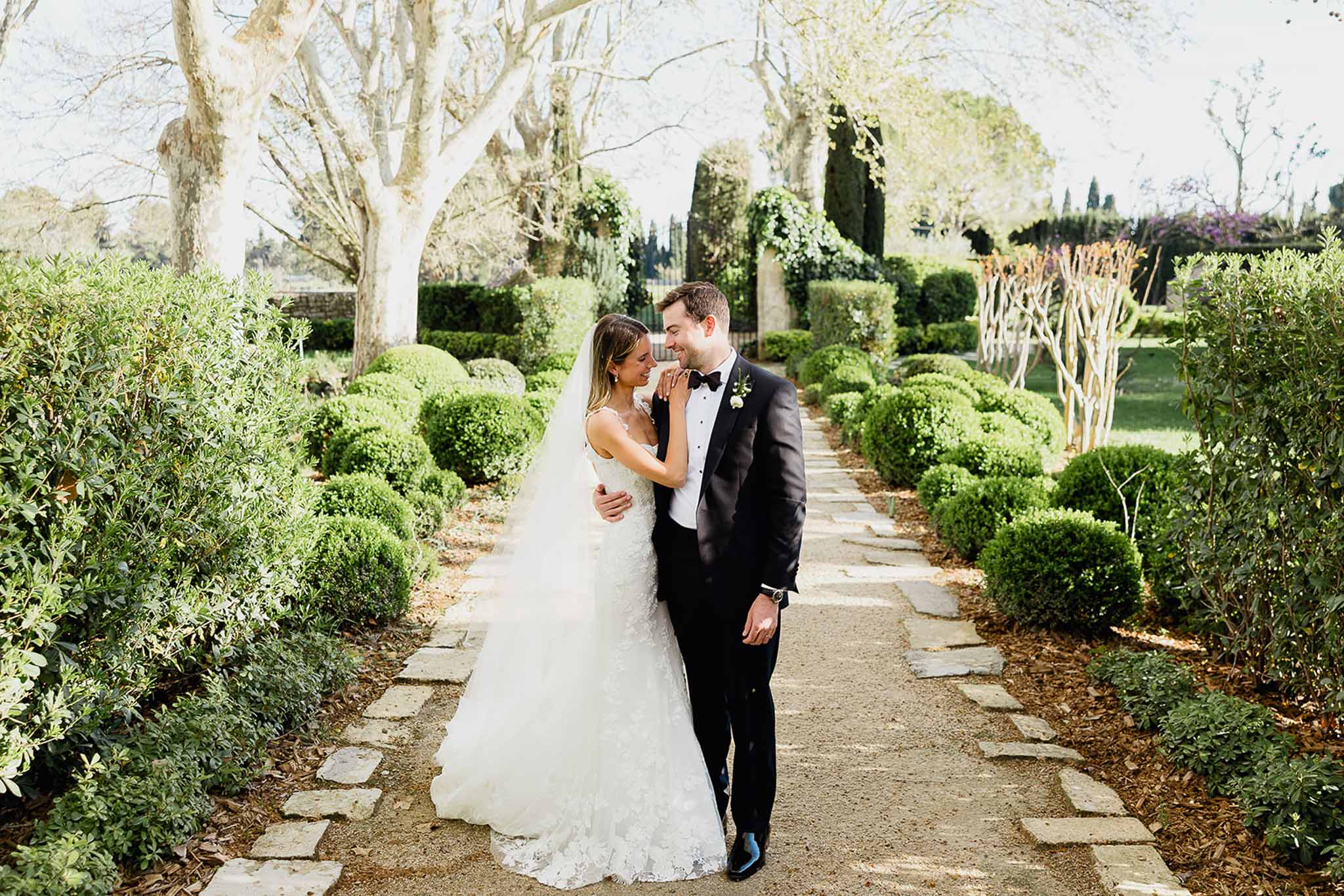 Bride and groom walking down stone pathway in formal garden with topiary hedges and mature trees