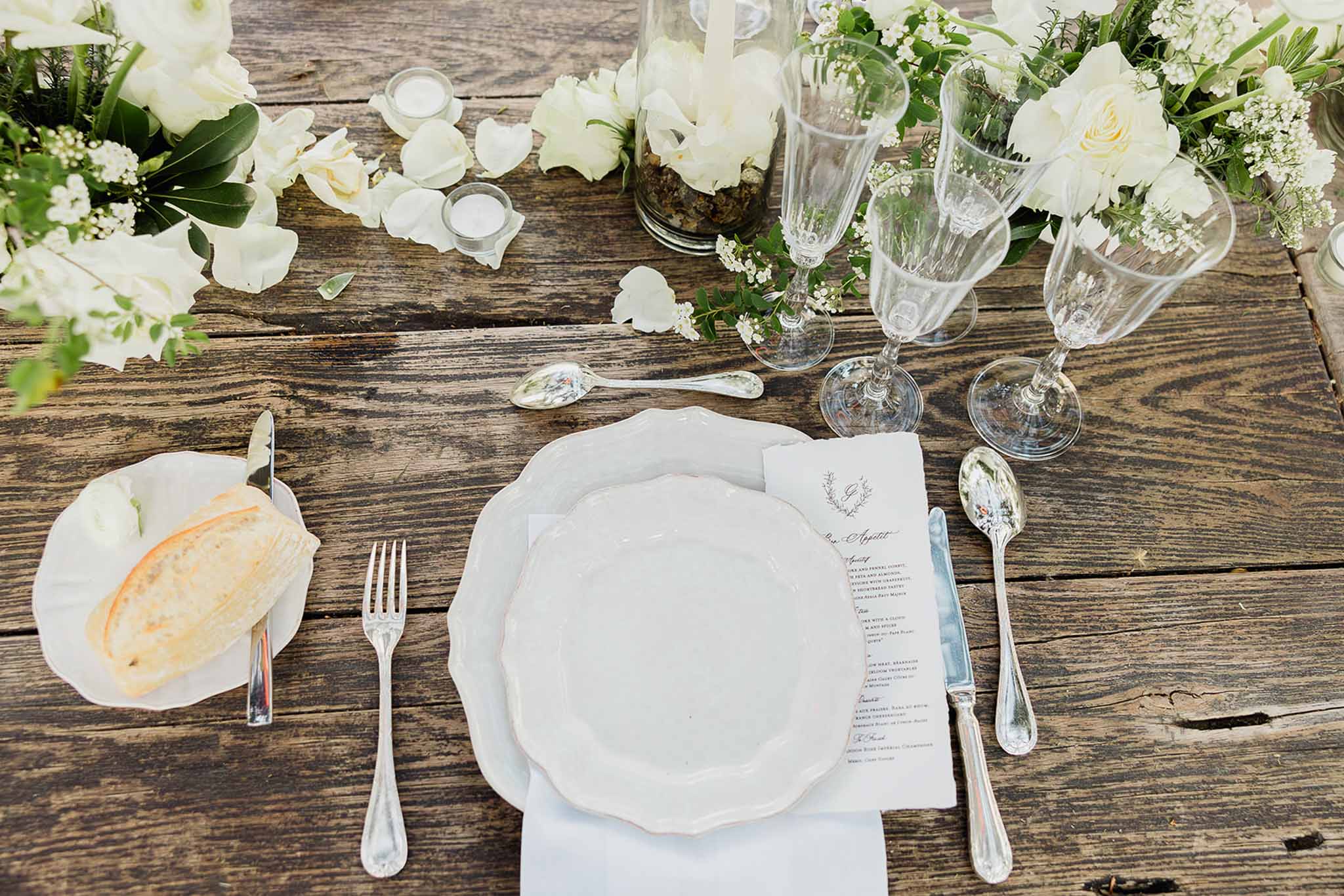Overhead view of rustic wedding table setting with white plates, silverware and cream roses on dark wood table