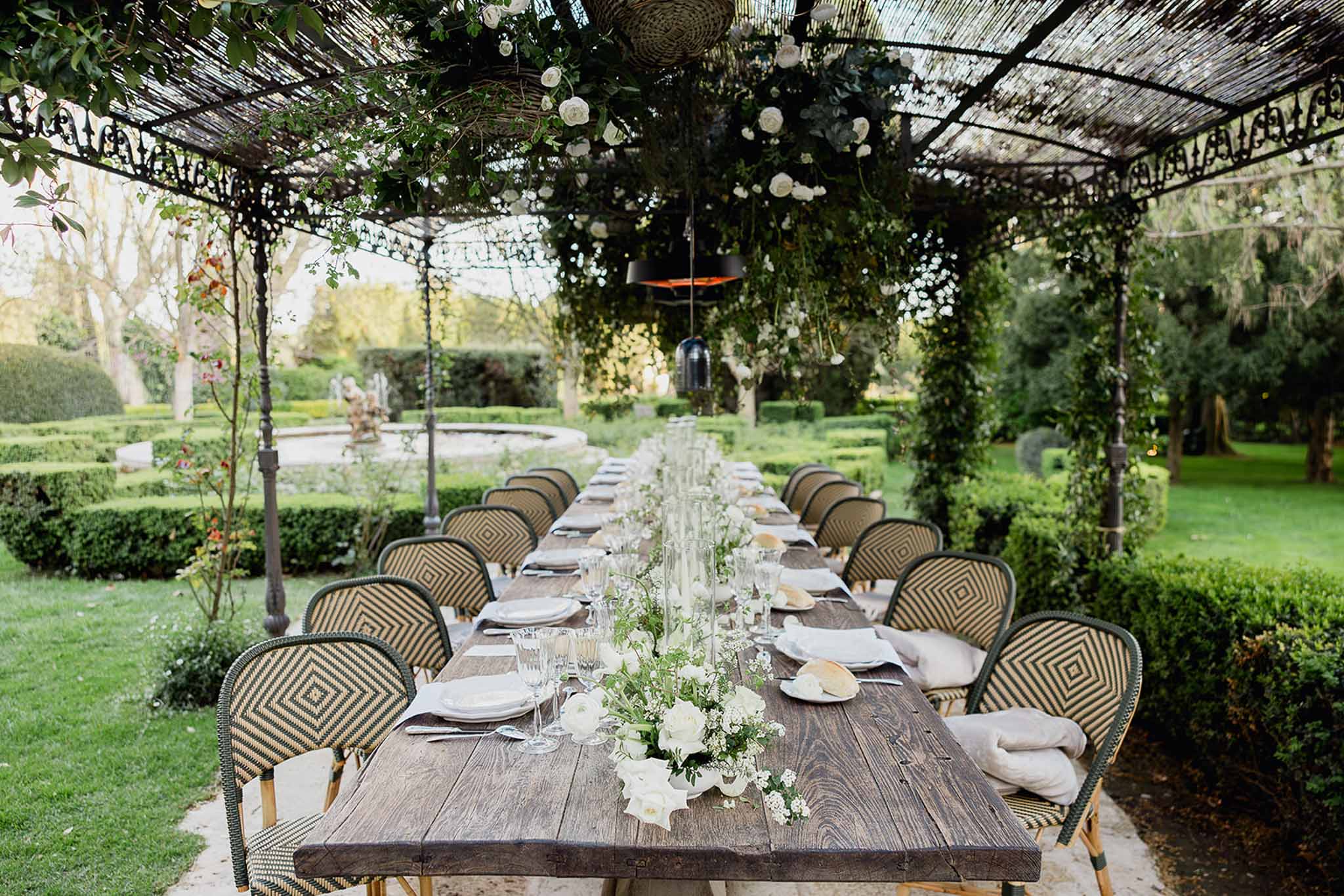 Outdoor reception table with floral arrangements in formal garden with fountain and pergola