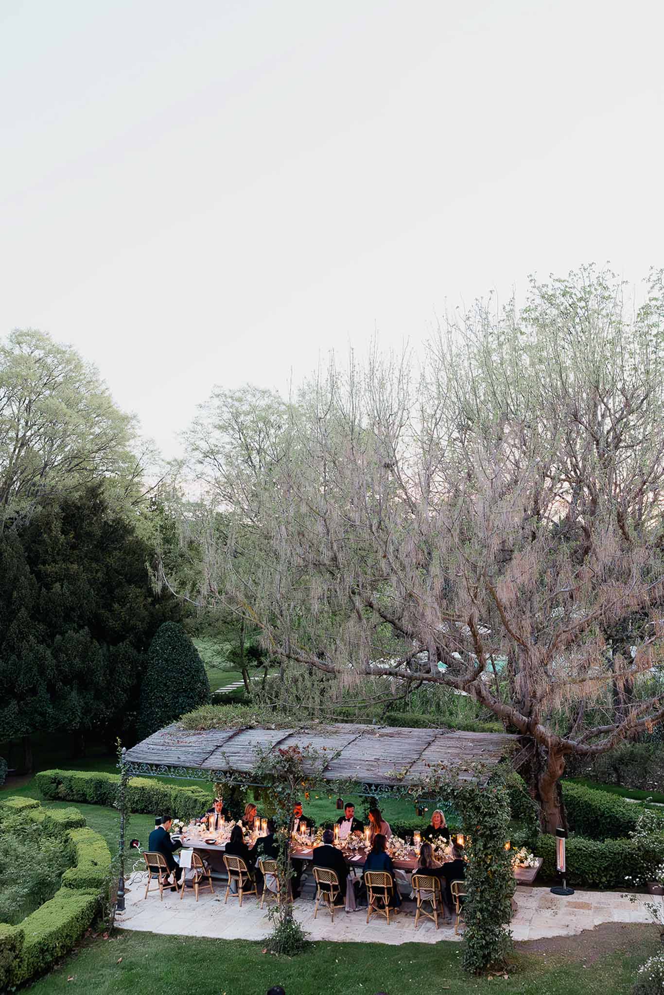 Outdoor wedding reception dinner under pergola in formal garden with guests at long table