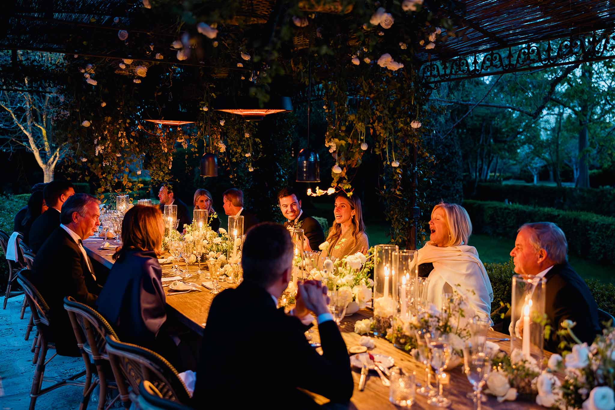 Evening wedding reception dinner with guests at long table under decorated pergola with lanterns and greenery