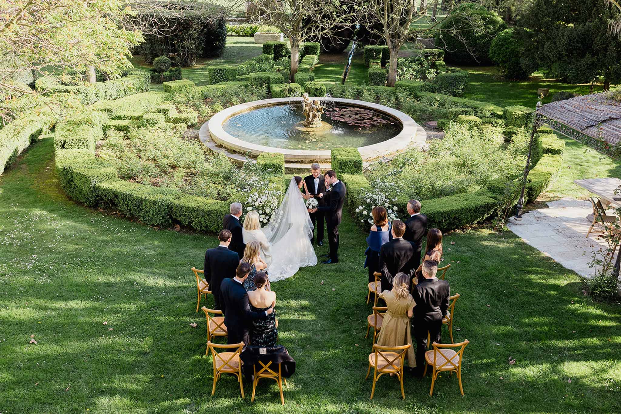 Aerial view of outdoor wedding ceremony in formal garden with stone fountain and manicured hedges
