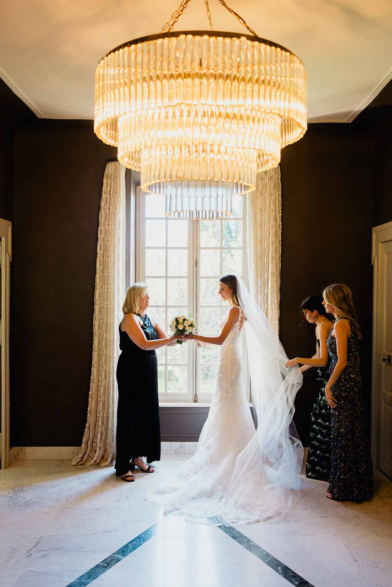 Bride with mother and bridesmaids getting ready indoors beneath elegant chandelier