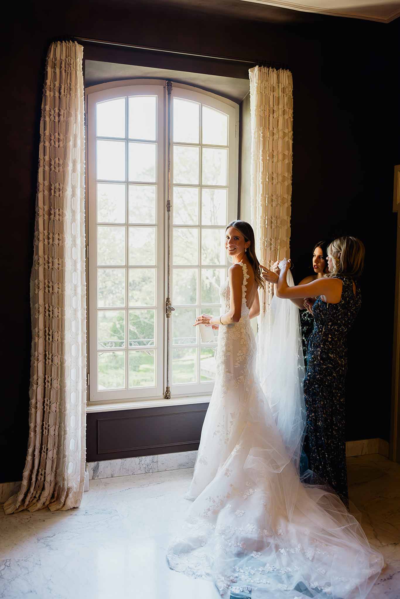Bride being dressed by attendants in elegant indoor getting ready room with French doors