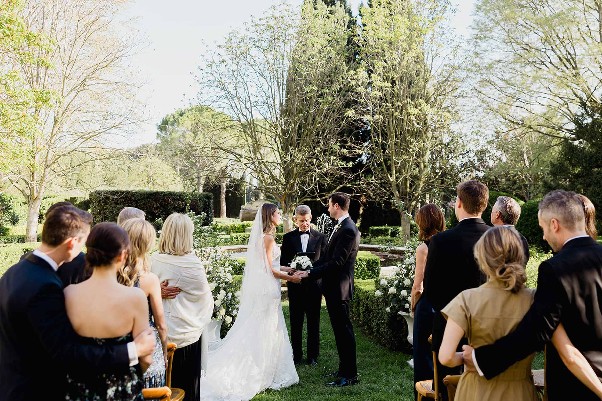 Outdoor wedding ceremony with bride and groom exchanging vows in formal garden setting with guests