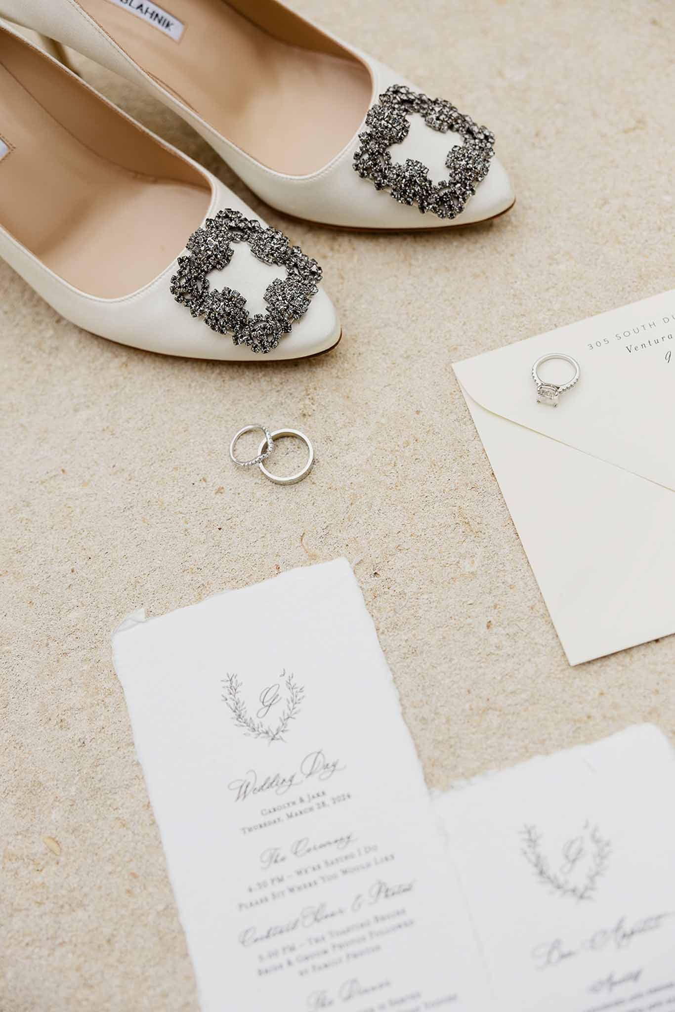 Wedding flat lay with ivory bridal shoes, silver rings, and white programs on stone surface