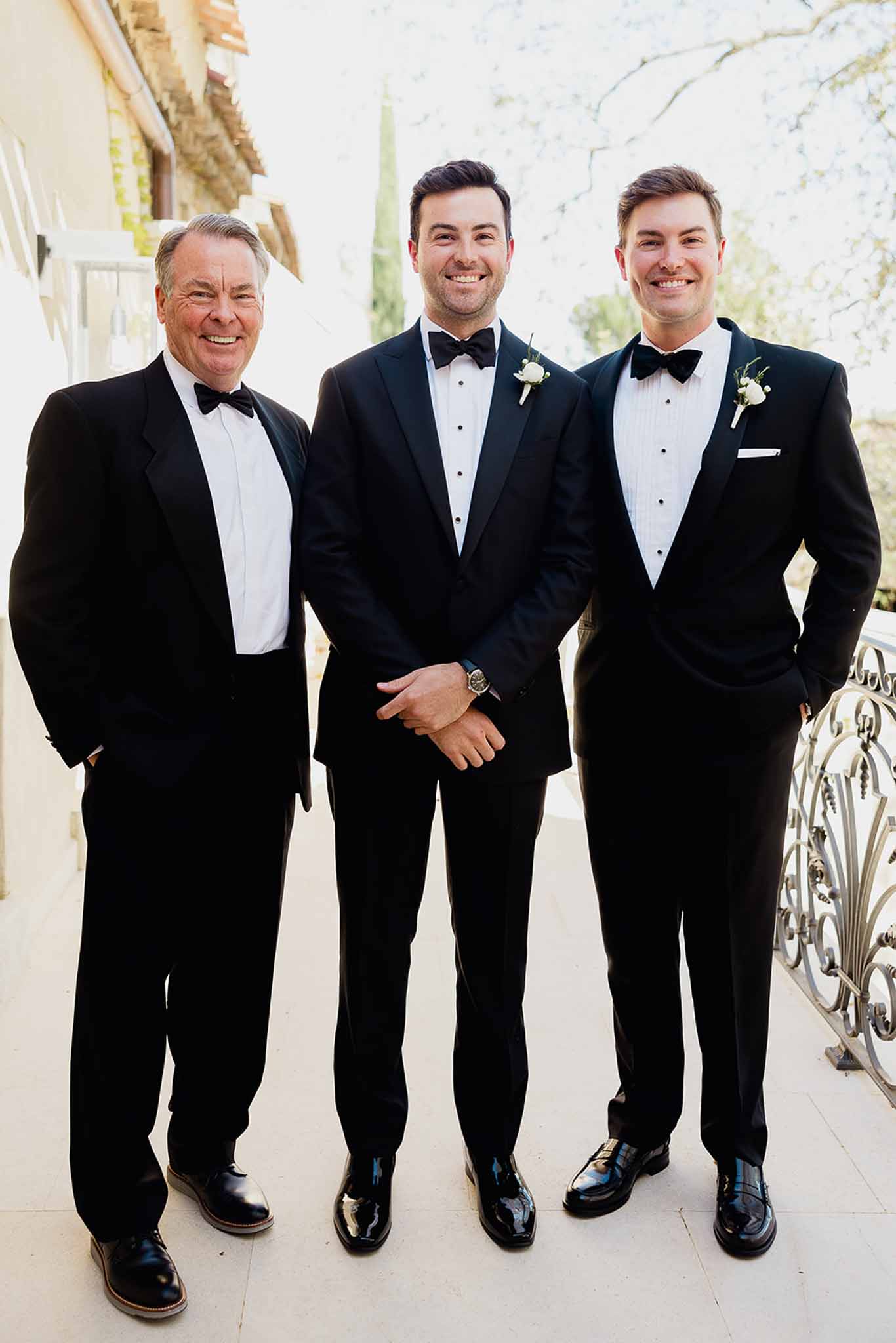 Groom with two groomsmen in black tuxedos on stone terrace at elegant wedding venue