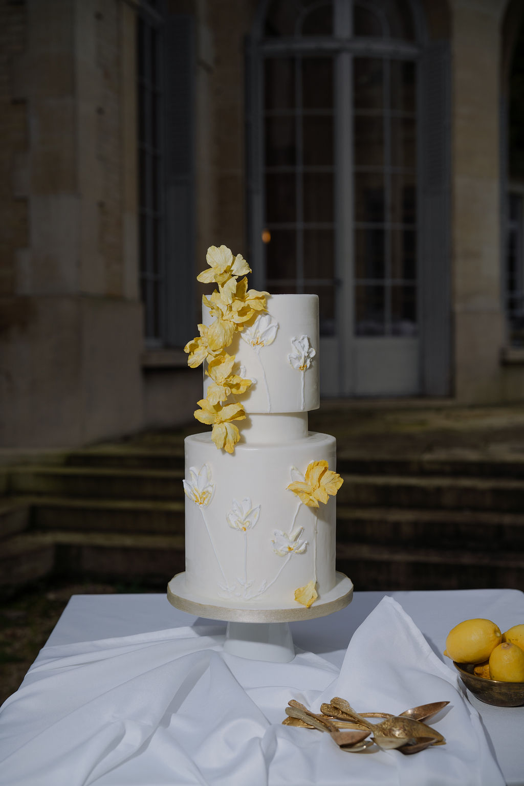 Two-tiered white wedding cake with yellow sugar flowers and lemons on linen table before a chateau