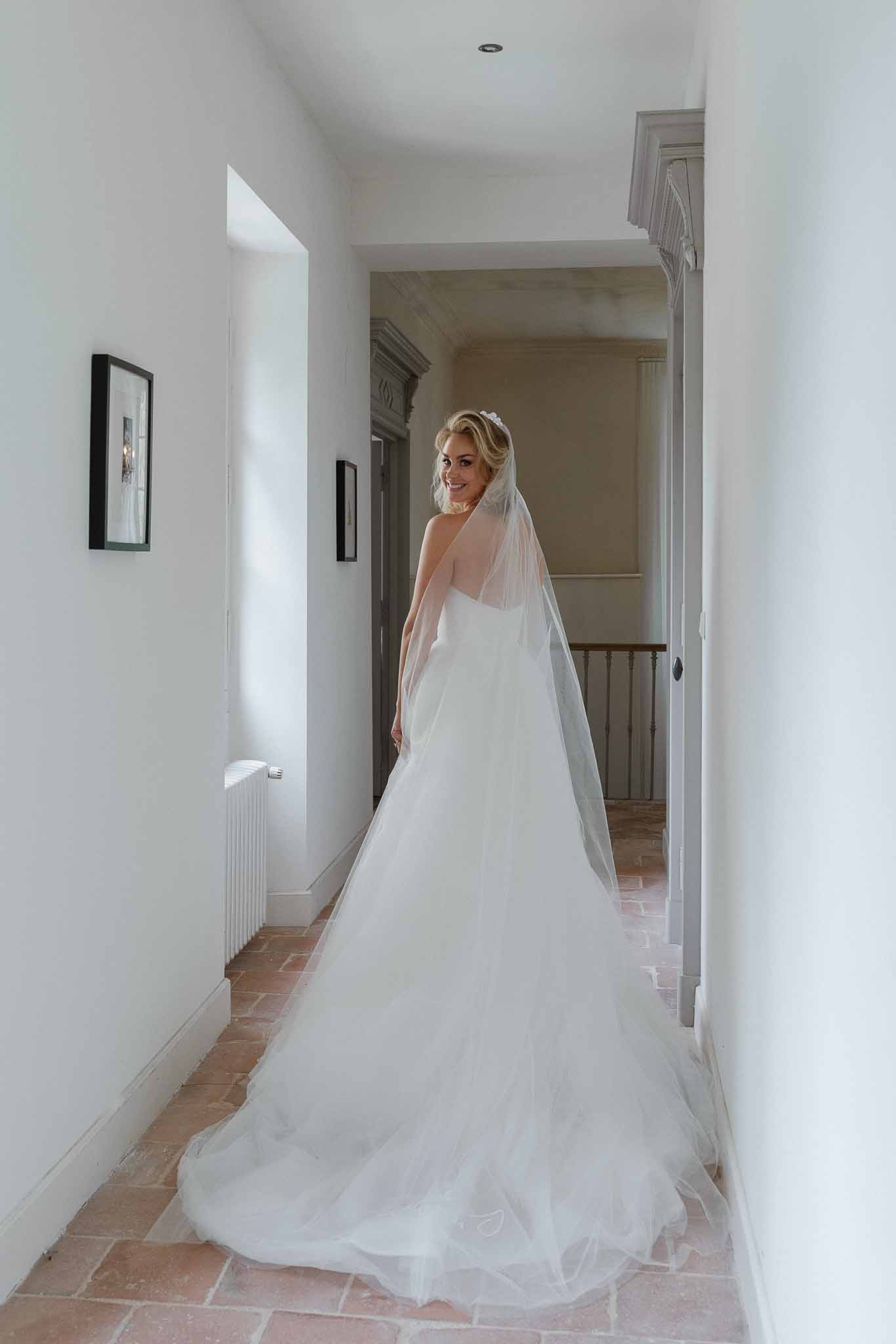 Bride in ivory gown and veil walking down elegant hallway with terracotta floors